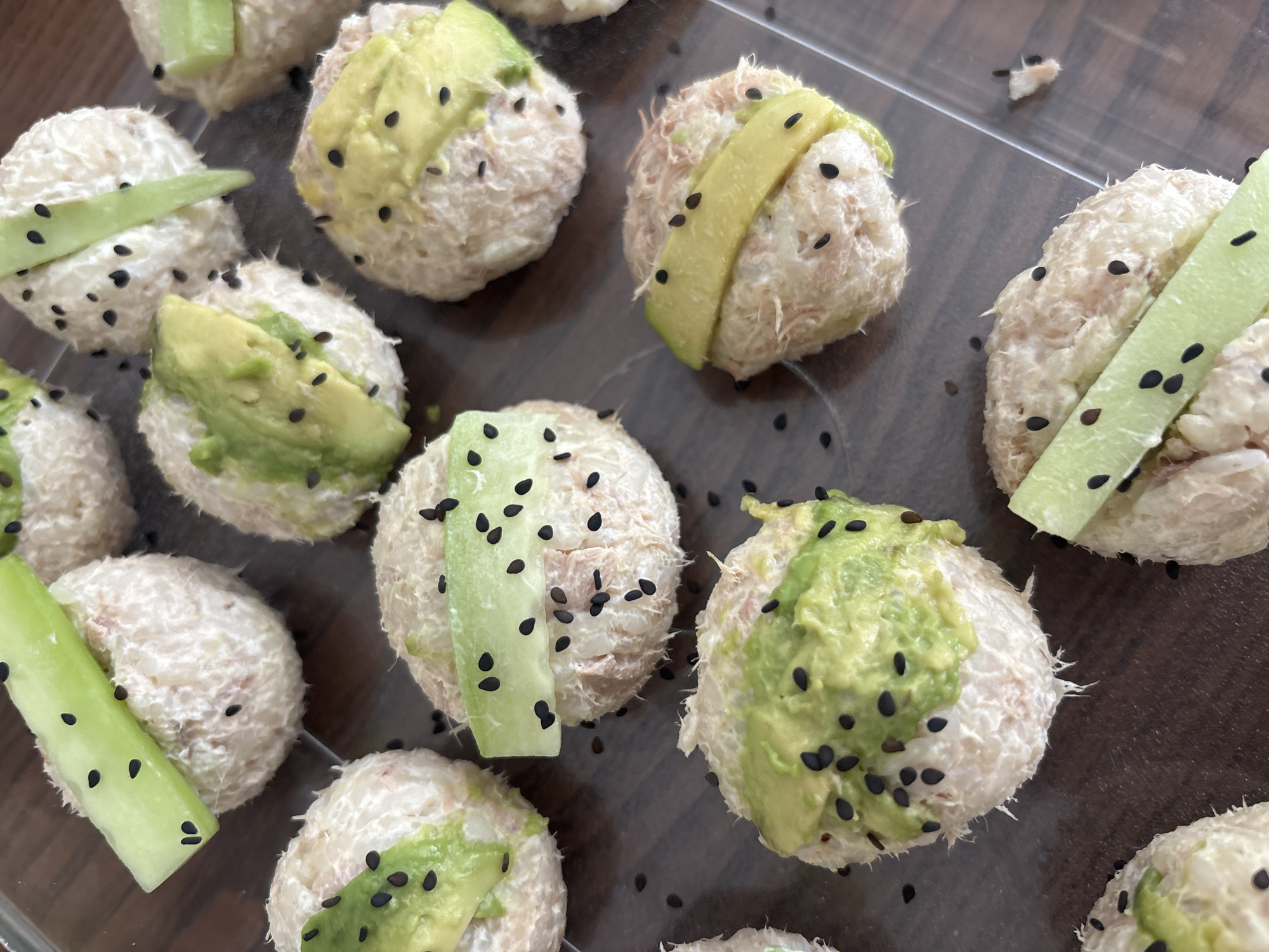 A close-up photograph displays a collection of sushi-like appetizers arranged on a clear, plastic tray. The tray is placed on a brown surface. The appetizers are round and appear to be made of a light-colored, shredded filling coated in black sesame seeds. Some are topped with sliced avocado or cucumber. The location in Puebla, Mexico is unknown.