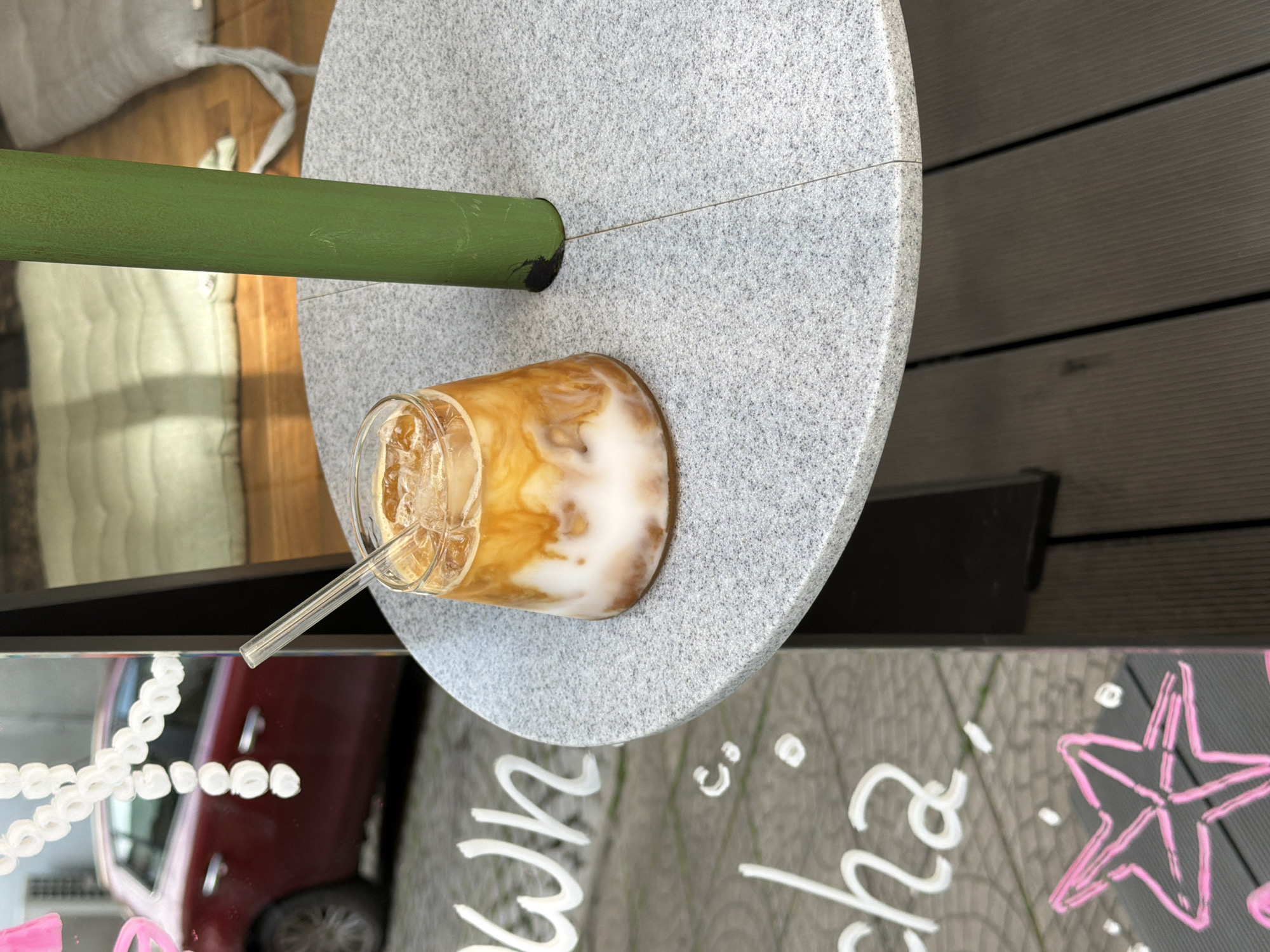 The image shows an iced coffee drink with a straw in a clear glass, placed on a speckled gray table. The drink appears to be a mix of coffee and milk or cream, creating a marbled effect. The table has a smooth, circular top and is supported by a thick green cylindrical leg. In the background, wooden planks are visible, suggesting an outdoor setting. A glimpse of a red car is reflected in a mirrored surface in the foreground, which also has white and pink chalk drawings of stars and text. The text appears to say "cha" and "wn" with other illegible words. The lighting suggests it is daytime, likely sunny. The overall scene appears to be at a cafe or an outdoor seating area.