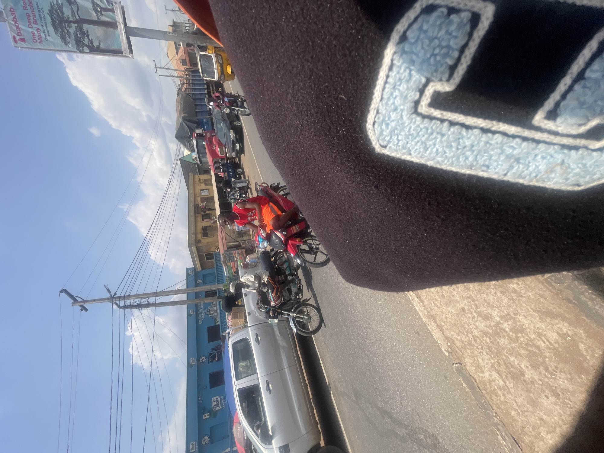 The image depicts a busy street scene in Lokoja, Nigeria. A variety of vehicles, including motorcycles, cars, and a truck, are present. There are individuals riding the motorcycles and occupying the vehicles. A blue building and a billboard are visible in the background. The image appears to be taken from a slightly elevated perspective, possibly from inside a vehicle or building.