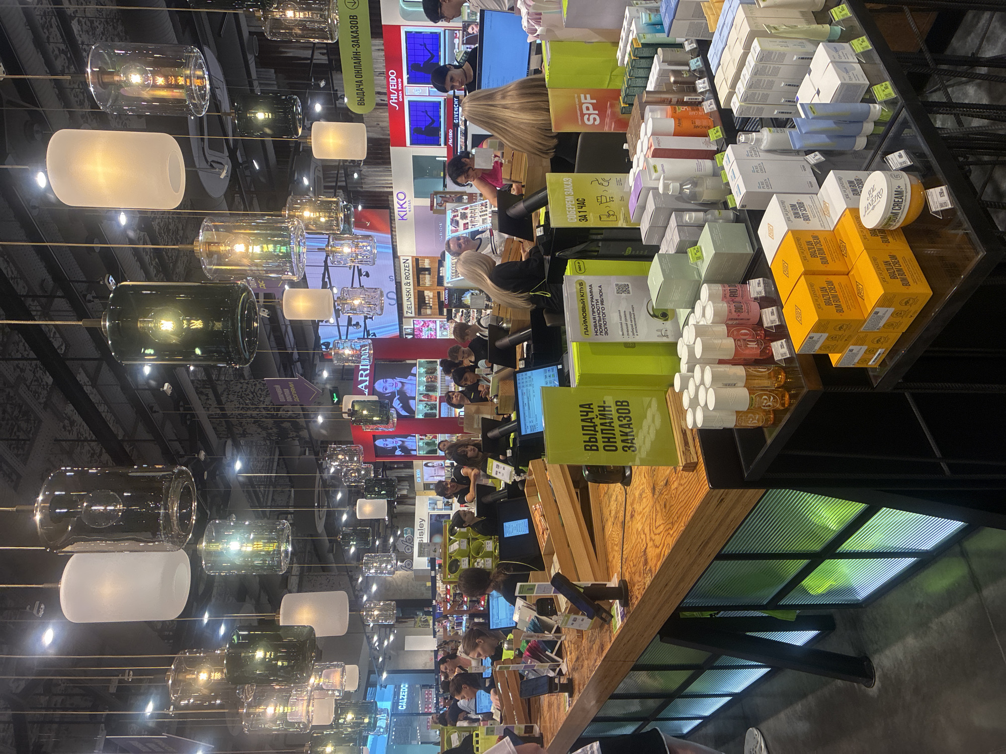 The image depicts an interior view of a retail store, likely a cosmetics or personal care shop, illuminated by numerous hanging pendant lights. The store appears to be well-stocked, with shelves and display tables filled with various products, including boxes, bottles, and tubes. Several people are visible in the scene, some working behind a counter with computer monitors, and others browsing or interacting with staff. The counter area features wooden surfaces and a bright green sign with Cyrillic text that appears to indicate "Online Order Pickup." In the background, brand names like "SHISEIDO," "KIKO MILANO," and "CALZEDONIA" are discernible, suggesting a multi-brand retail environment. The overall atmosphere is that of a busy commercial space. Without explicit signage or context within the image itself, pinpointing the exact location within Sochi, Russia, is not possible based solely on the visual information provided.