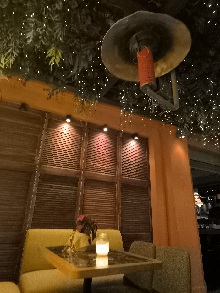 This image depicts a warm and inviting interior or semi-outdoor seating area, characteristic of a restaurant, bar, or cafe, likely in a location such as Palaio Faliro, Greece. The scene is set during the evening or night, illuminated by various artificial light sources.
The ceiling is adorned with lush artificial greenery, densely woven with numerous small, twinkling string lights that create a festive and magical ambiance. A large, sombrero-shaped outdoor heater with a glowing red heating element hangs prominently from the ceiling, suggesting the space might be open-air or requires supplemental warmth.
In the foreground, a small square table with a dark, reflective surface is positioned between two upholstered seating options: a mustard-yellow booth in the immediate foreground and another partially visible behind the table. A small potted plant with reddish-brown leaves and a delicate candle flickering in a glass holder grace the tabletop, adding a cozy detail. Part of a textured green armchair is visible to the right of the table.
The walls feature a mix of materials and colors. The background is dominated by warm brown wooden panels, possibly decorative shutters or slatted screens, which are softly illuminated by three recessed spotlights casting reddish hues. An upright pillar and a section of the wall on the right are painted in a vibrant orange or terracotta shade, contributing to the Mediterranean aesthetic. Blurred elements in the deeper background suggest other parts of the establishment or an outdoor view. No people are visible, emphasizing the serene and prepared atmosphere. There is no visible text in the image.