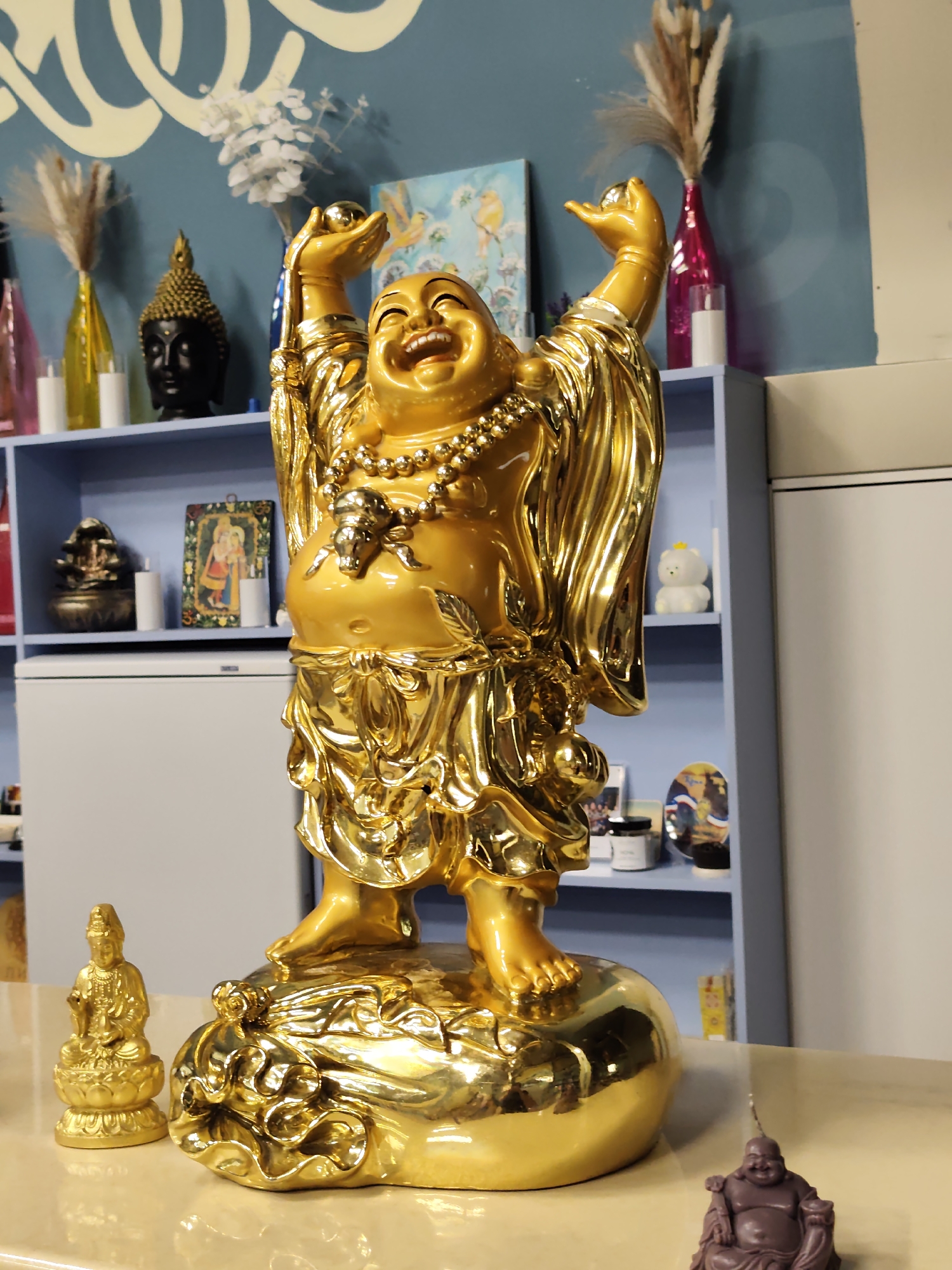 The image depicts a collection of Buddhist statues and decorative items on a table and shelves against a blue wall with a white mural. Dominating the foreground is a large, golden statue of a laughing Buddha with arms raised and holding a ball, placed on a golden sack. To the left of the large statue is a smaller, seated golden statue of what appears to be Guan Yin. In the background, on blue shelves, are various items including a black Buddha head, decorative vases with dried flowers, a small framed image, a miniature stupa-like object, and a white bear-shaped figurine. The environment appears to be an indoor setting, possibly a shop or a home. No people are visible in the image. The provided information about the location being Sochi, Russia, cannot be verified from the image content itself.