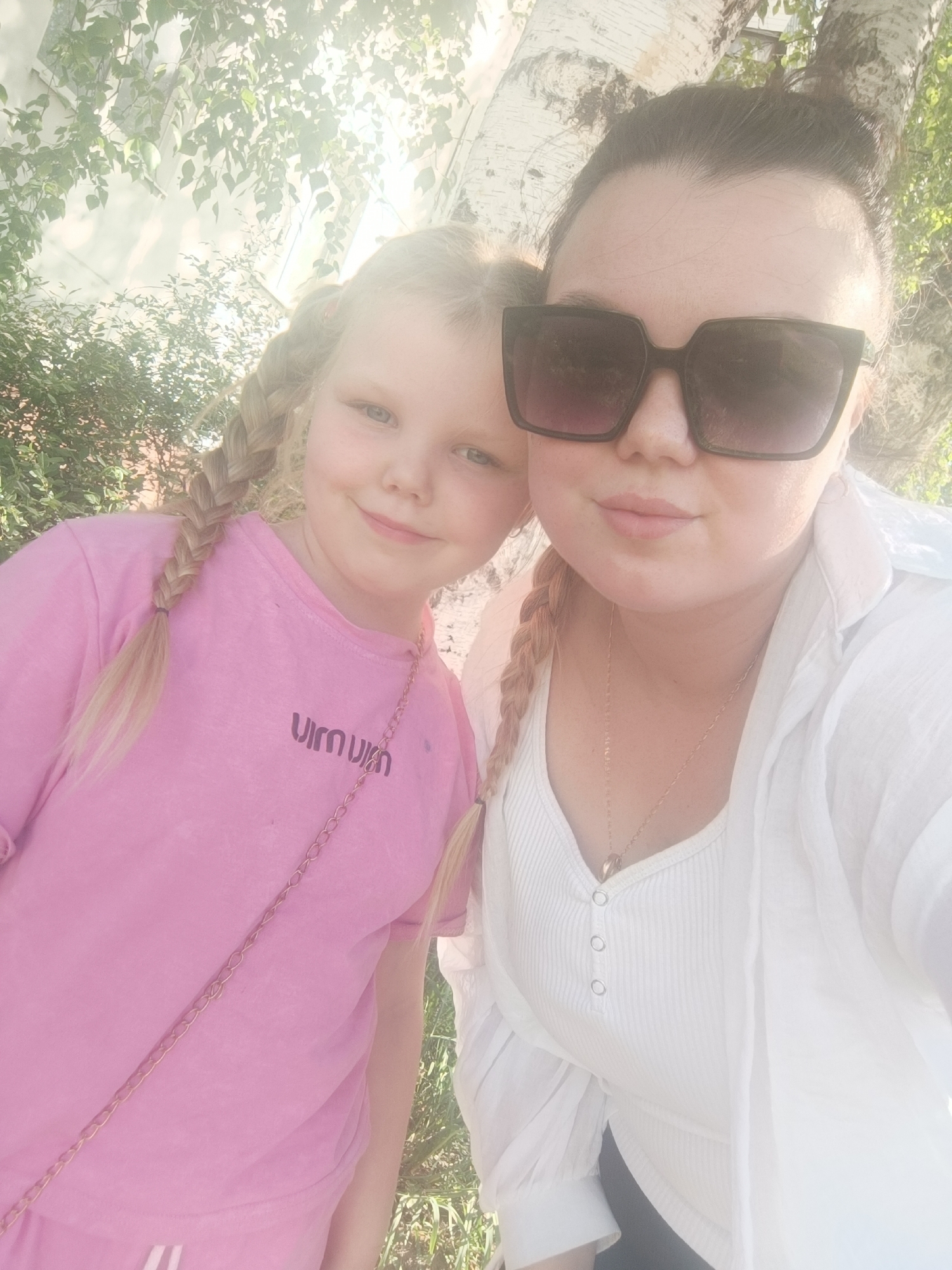 Two females are in the image, likely a young girl and an adult woman. The girl, on the left, has light-colored hair styled into pigtail braids. She is wearing a pink T-shirt with a chain strap across her body. The woman, on the right, wears large, dark sunglasses and also has her hair in braids. She wears a white shirt and a gold necklace. The location appears to be outdoors, with greenery and a white building visible in the background. The image has bright sunlight, which may suggest a day time shot. Unknown, Unknown city is not identifiable in the picture.