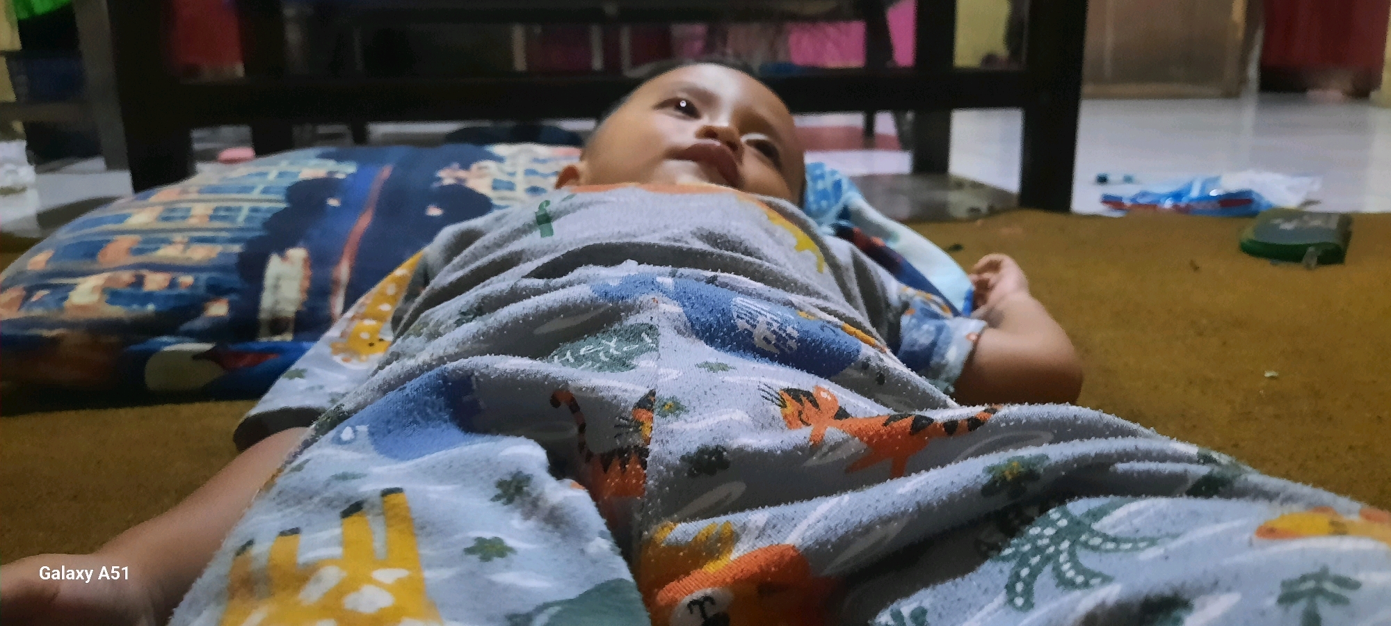 In Jatitengah, Indonesia, a low-angle shot captures a close-up of a baby lying on its back. The baby is wearing pajamas with a cartoon animal print featuring giraffes and tigers. A blue patterned pillow is visible beneath the baby's head. The environment appears to be indoors, with a brown carpeted floor and dark furniture in the background. The image is watermarked with "Galaxy A51."