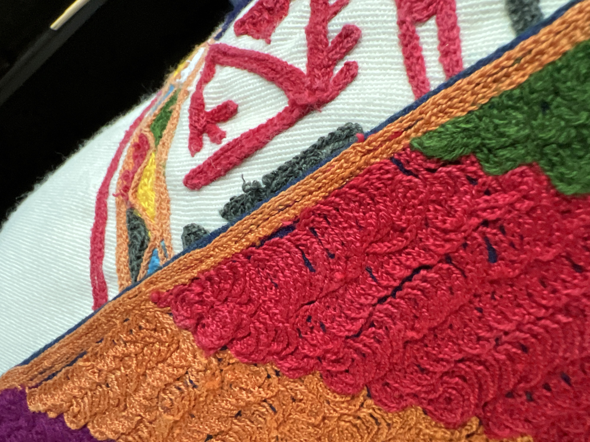 The image displays a close-up, oblique view of a vibrantly colored textile, likely a piece of embroidery or crochet work. The fabric is composed of multiple distinct sections with varied textures and patterns.
The dominant portion of the image on the left features large, textured blocks of color. A prominent area in the lower-left to mid-left is a dense, raised pattern of bright red yarn, appearing as clusters of loops or stitches, creating an almost scalloped or berry-like texture. Adjacent to this, in the upper-left, is a similar but distinct pattern in bright orange yarn, exhibiting a tightly packed, woven or crocheted texture with visible stitches. A smaller section of deep purple is partially visible in the far upper-left corner. Separating these textured blocks from a lighter background is a narrow, braided or tightly stitched border of orange yarn, frequently outlined by a thin line of dark blue thread. A section of dark green, similarly textured yarn, is visible in the lower-right of the red block.
The right side of the image reveals a white or off-white woven background fabric, characterized by horizontal lines indicating its weave structure. On this white background, various abstract or geometric patterns are stitched in contrasting colors. These include thin, linear designs in pink, yellow, green, orange, and gray. Several prominent red, thick, linear stitches form angular, branching, or zig-zagging shapes against the white, reminiscent of stylized letters or symbols. Some of these red lines appear to form inverted "V" or "Y" shapes.
The overall texture of the colored sections is three-dimensional and tactile, contrasting with the flatter, woven background. The light source appears to be from the upper left, highlighting the raised textures. A dark, out-of-focus area is visible in the upper-right corner, suggesting a background behind the textile, but no specific objects are discernible there. There are no people, actions, or interactions depicted in the scene.