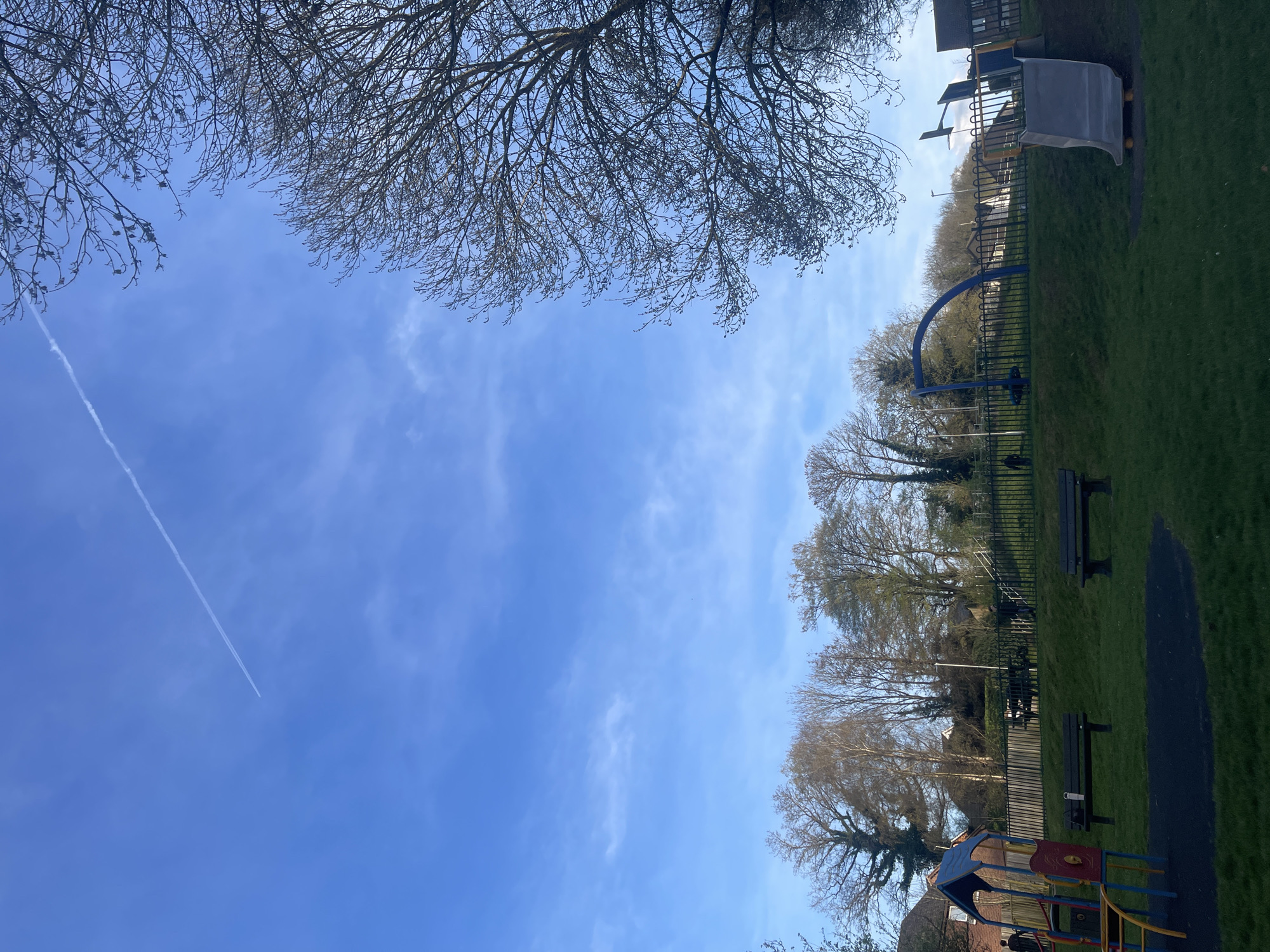 The image shows a park in Maidstone, United Kingdom, on a sunny day. The sky is blue with sparse cirrus clouds and a contrail. A large bare tree is visible in the upper left corner. On the right side, a grassy area slopes upwards, containing park benches, playground equipment, and trees. A fence runs along the edge of the park. A partially visible playground structure is in the lower right corner. No people are visible.