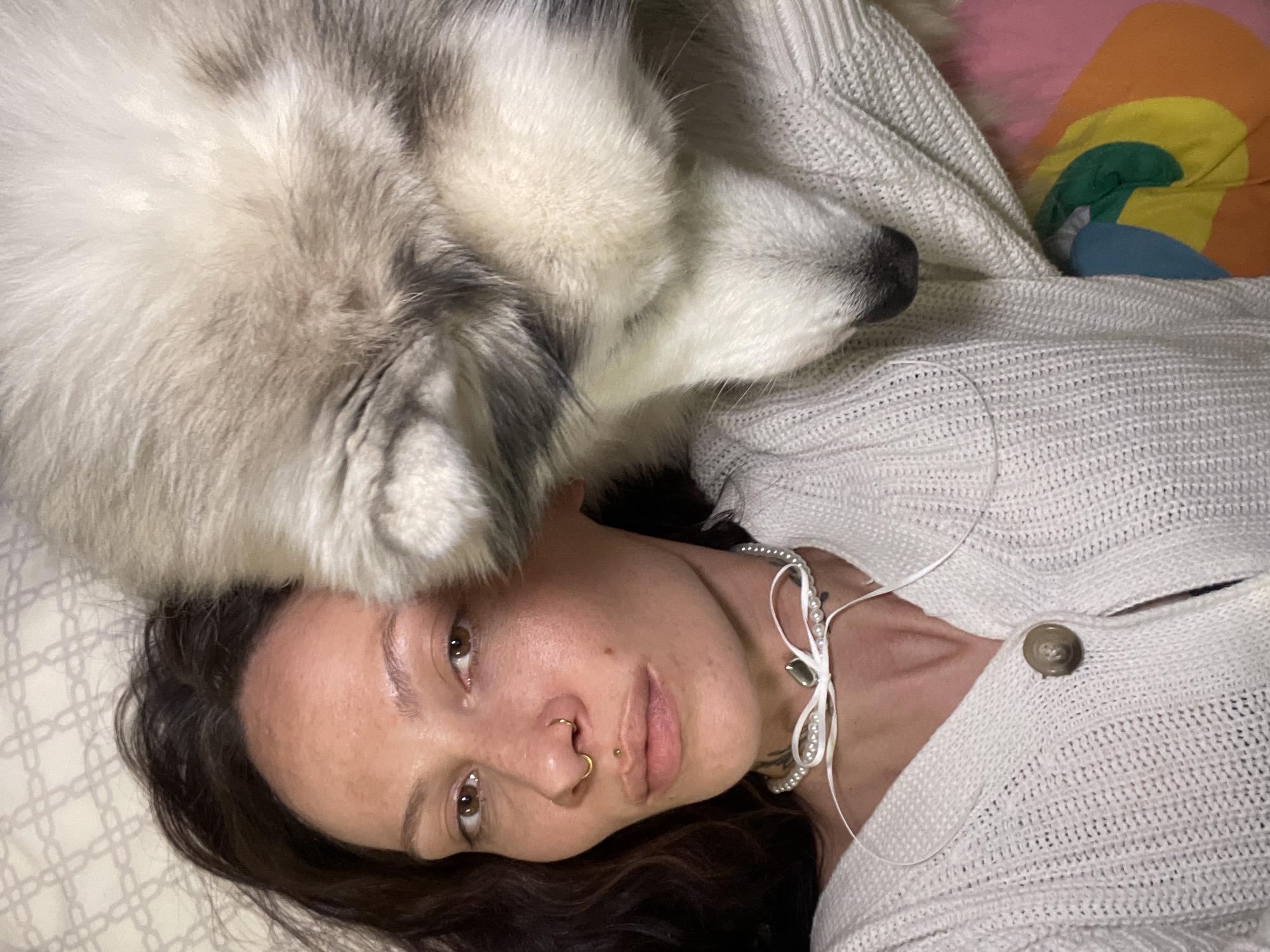 The image displays a close-up, high-angle shot of a woman and a dog resting intimately, likely indoors in Izhevsk, Russia.
The primary subject is a woman lying on her back, looking directly at the viewer with a calm, neutral expression. She has long, dark hair spread out beneath her head and features light skin and brown eyes. Notable details on her face include a gold hoop piercing in her left nostril and a small gold stud above her upper lip. She is wearing a light-colored, possibly off-white or cream, knitted cardigan with a single brown button visible on the right side of her chest. Around her neck, she wears a decorative necklace composed of white, pearl-like beads strung together with delicate white ribbons.
Beside and above the woman's head is a fluffy, grey and white dog, resembling a husky or similar breed, also lying down. Its voluminous fur, a mix of white and various shades of grey, covers a significant portion of the upper frame, and its nose is visible on the far right. The dog appears to be snuggled close to the woman, suggesting companionship.
Both the woman and the dog are resting on a light-colored, possibly white, patterned bedsheet or blanket. In the background, behind the dog and the woman's shoulder, a colorful pillow featuring segments of orange, yellow, green, and blue is partially visible. The scene conveys a moment of quiet relaxation and connection between the woman and her pet. No visible text is present in the image.