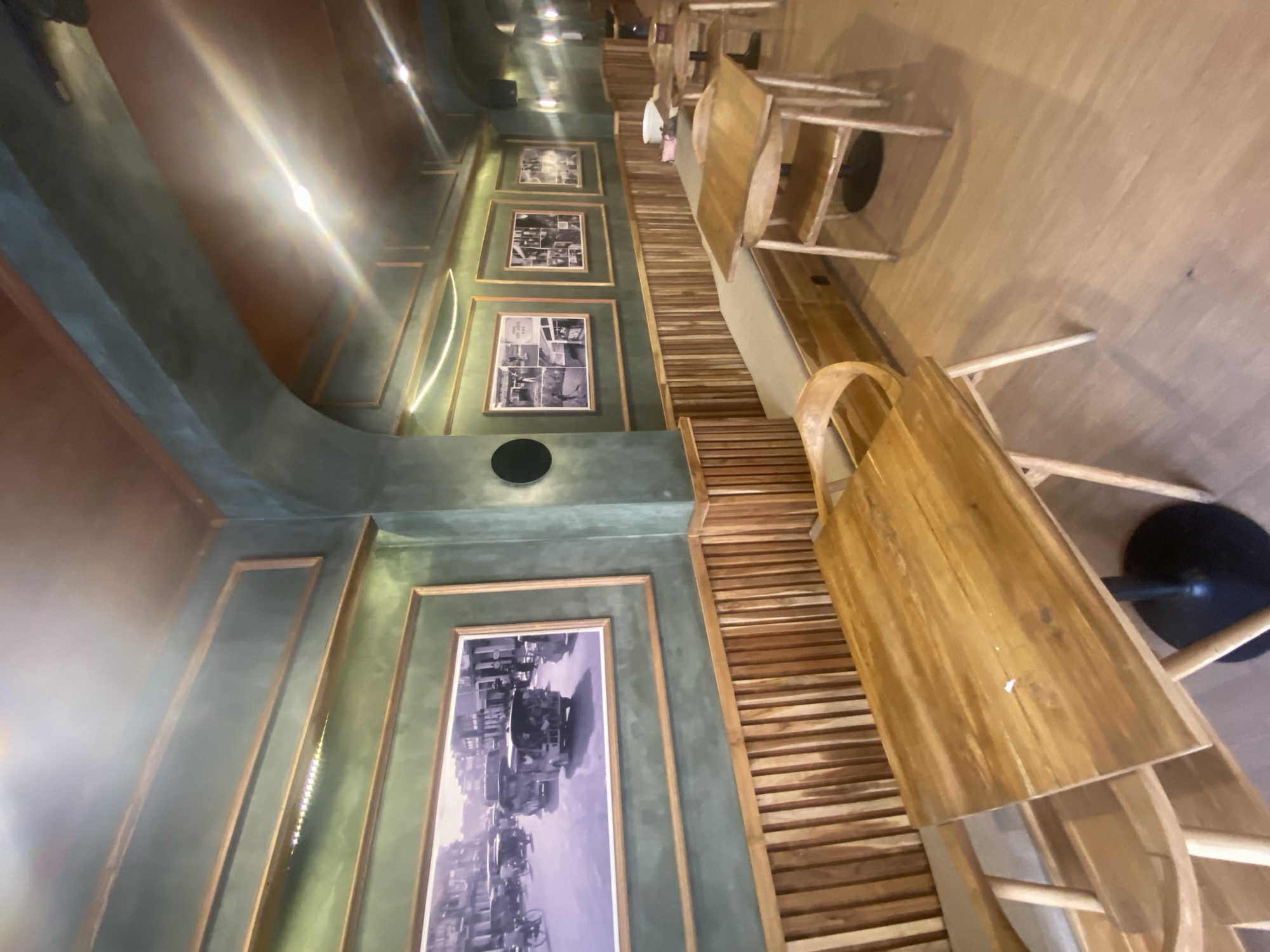 The image depicts the interior of a cafe located in Denpasar, Indonesia. The cafe features a rustic and vintage ambiance, with walls painted in a muted green hue and adorned with framed black and white photographs. The furniture is primarily made of light-colored wood, including tables and chairs. A long, upholstered bench runs along the wall, providing additional seating. The lighting appears to be warm and ambient, with some glare from overhead fixtures. The flooring is made of light wood planks. In the foreground, a wooden table with a black pedestal base is visible, and a wooden chair with a curved back is positioned beside it. Further into the cafe, other tables and chairs are arranged, some with a bench along the wall. The overall impression is of a cozy and stylish dining or lounging space.