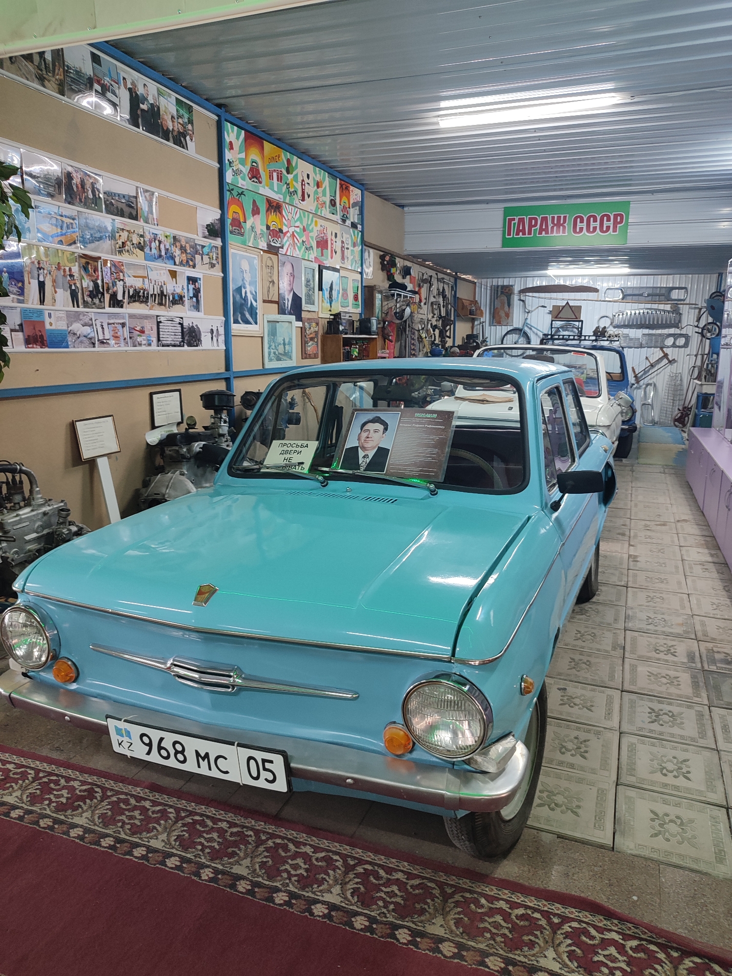 The image shows the interior of a garage museum in the city of Qonaev, Kazakhstan, labeled "GARAGE USSR" in Russian. The main subject is a light blue vintage car, possibly a ZAZ, positioned prominently in the foreground. The car has a license plate reading "KZ 968 MC 05." The interior of the garage is filled with various items, including engine parts displayed on the left, a collection of framed photographs and posters on the walls, and other vintage vehicles visible behind the main car. The walls are decorated with numerous framed pictures, some depicting groups of people, while others appear to be artwork or historical scenes. A bicycle is also visible in the background, along with various metal parts that might be car components or tools. The floor is tiled, and a patterned rug is placed in front of the main car. The overall impression is that of a museum dedicated to Soviet-era automobiles and memorabilia.