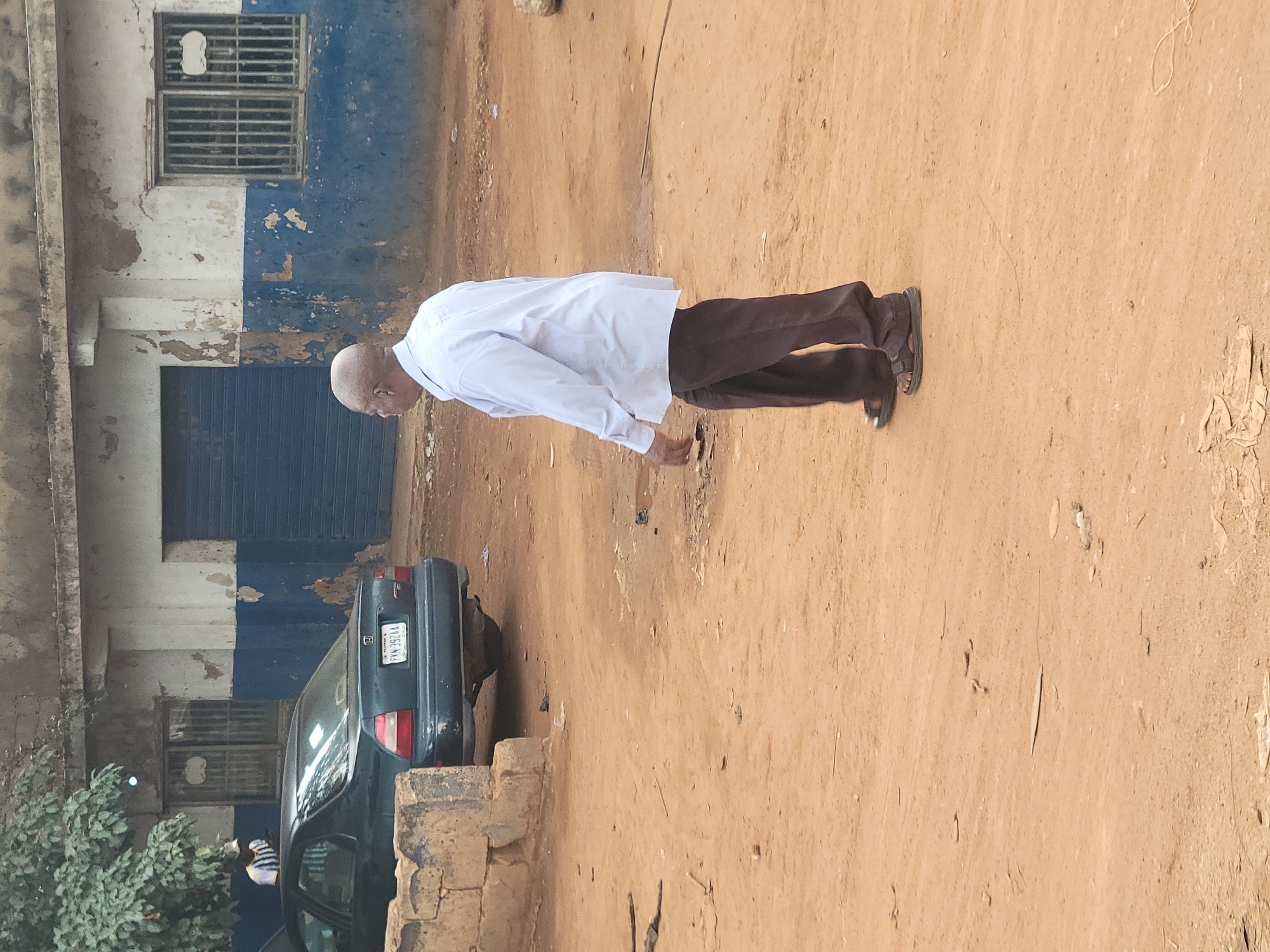 In Gura Top, Nigeria, a bald man in a light blue dress shirt and dark pants walks on a dirt path. He is wearing sandals. To his left, a dark green sedan is parked. Behind the car, a building with white and blue painted walls and barred windows is visible. A blue metal roll-up door is also present on the building. A person wearing a striped shirt and hat is partially visible near the car. The ground is covered in loose dirt and scattered debris.