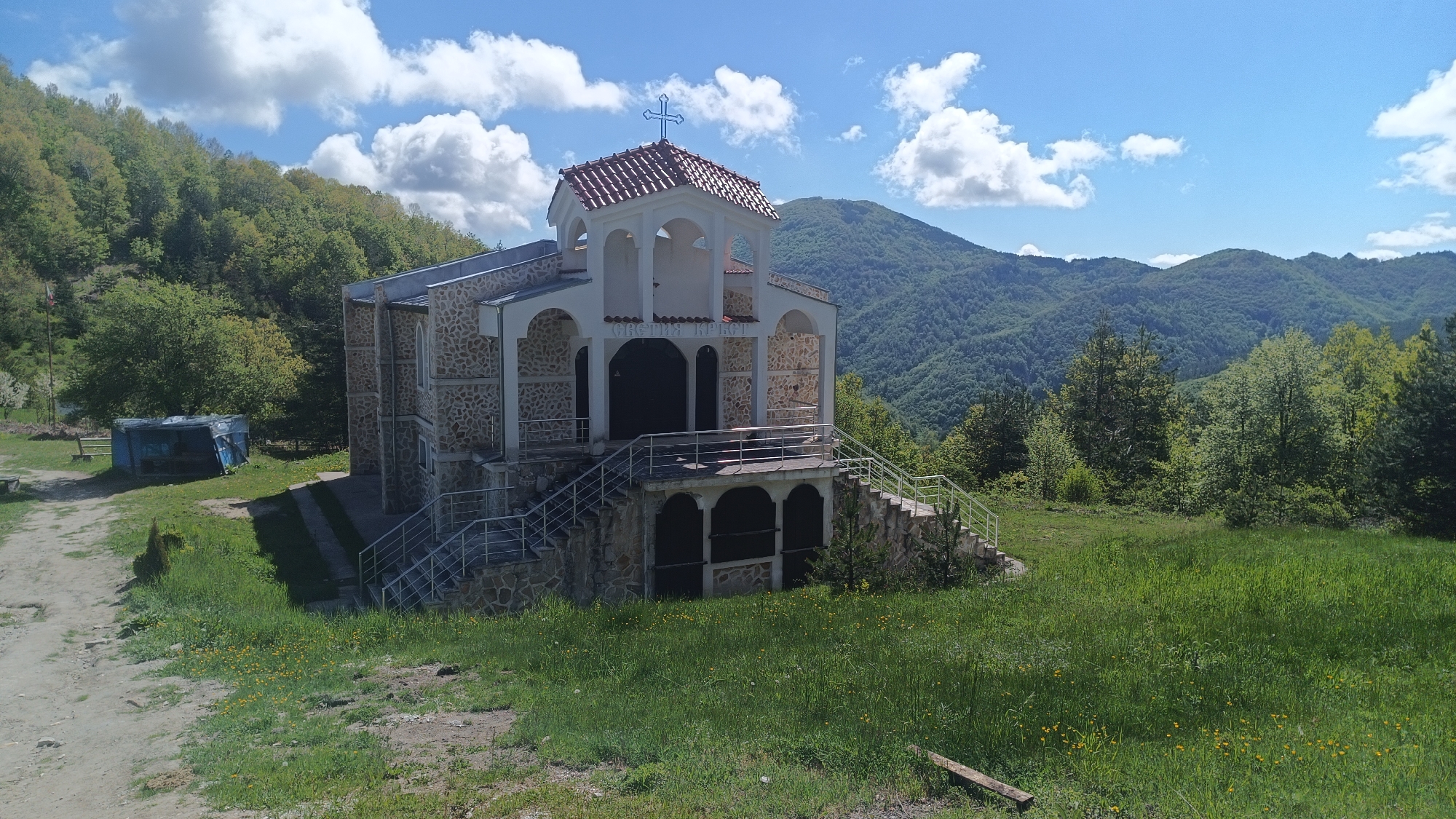The image depicts the Church of the Holy Cross in Sini Vrah, Bulgaria, a small structure built with stone and white architectural details, capped with a red tiled roof and a cross. The church is positioned on a grassy hillside with mountains visible in the background. A path or dirt road leads to the left of the church. Vegetation, including trees and grass, surrounds the church and slopes. The sky is blue with scattered clouds. A dilapidated blue shelter is also located in the vicinity of the church. No people are visible.