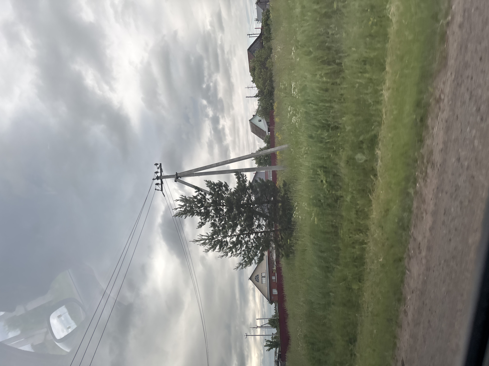 The image depicts a rural landscape under a cloudy sky, likely in the vicinity of Garevaya, Russia. The foreground shows a dirt road and a grassy field with scattered wildflowers. In the midground, a tall wooden power pole with insulators stands prominently, accompanied by power lines stretching across the frame. Behind the field, a red fence delineates a property line. A light-colored house with a triangular roof and arched windows is visible behind the fence, suggesting a residential area. Further back, several other houses and trees can be seen, along with more power poles and lines. The overall scene conveys a sense of rural tranquility, with the overcast sky contributing to a muted color palette.