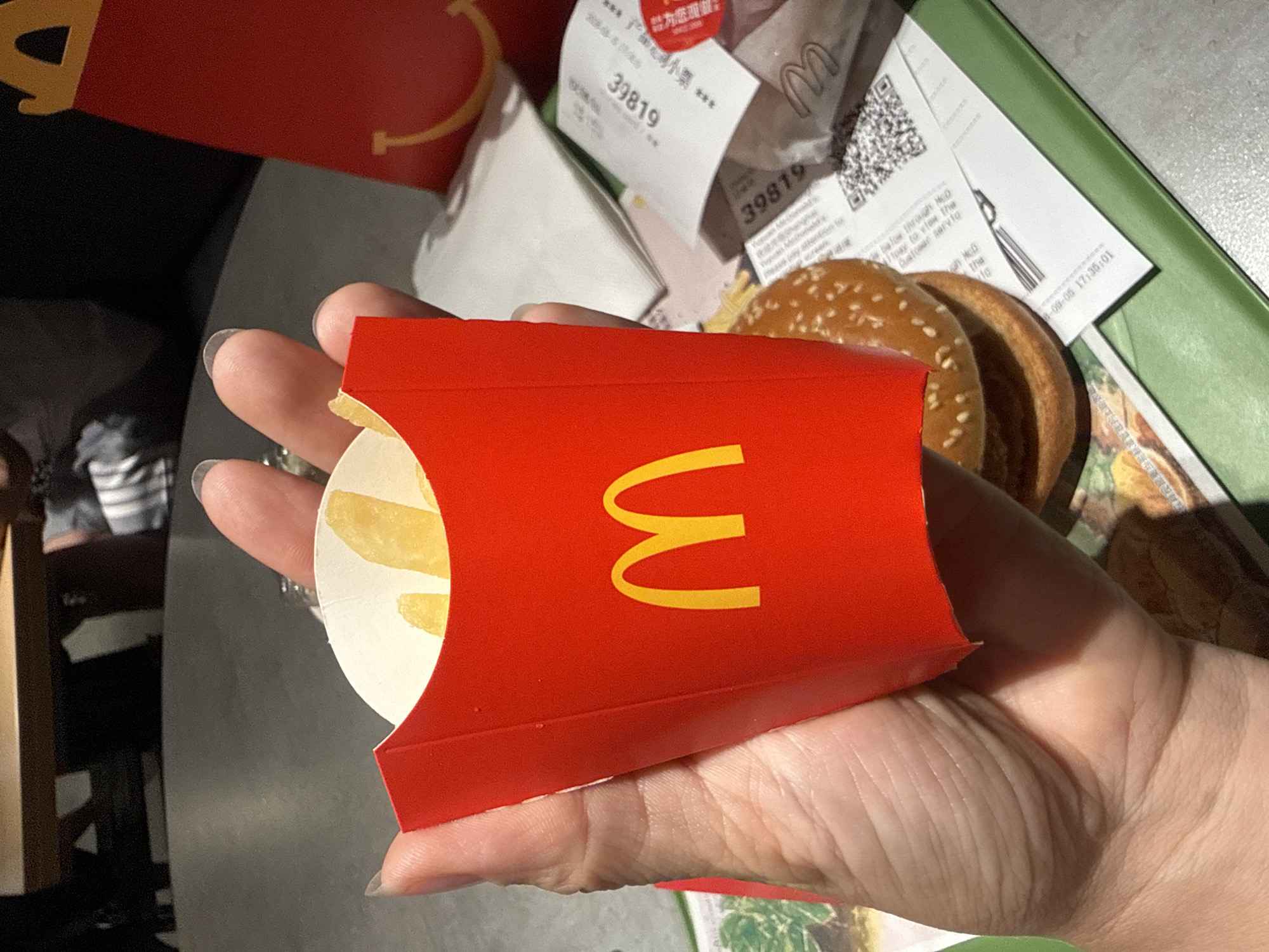 A hand is holding a red McDonald's french fry carton filled with french fries. The carton has the McDonald's logo on it. In the background, a McDonald's hamburger and receipts are visible on a green tray. The scene appears to be inside a McDonald's restaurant in Shanghai, China.