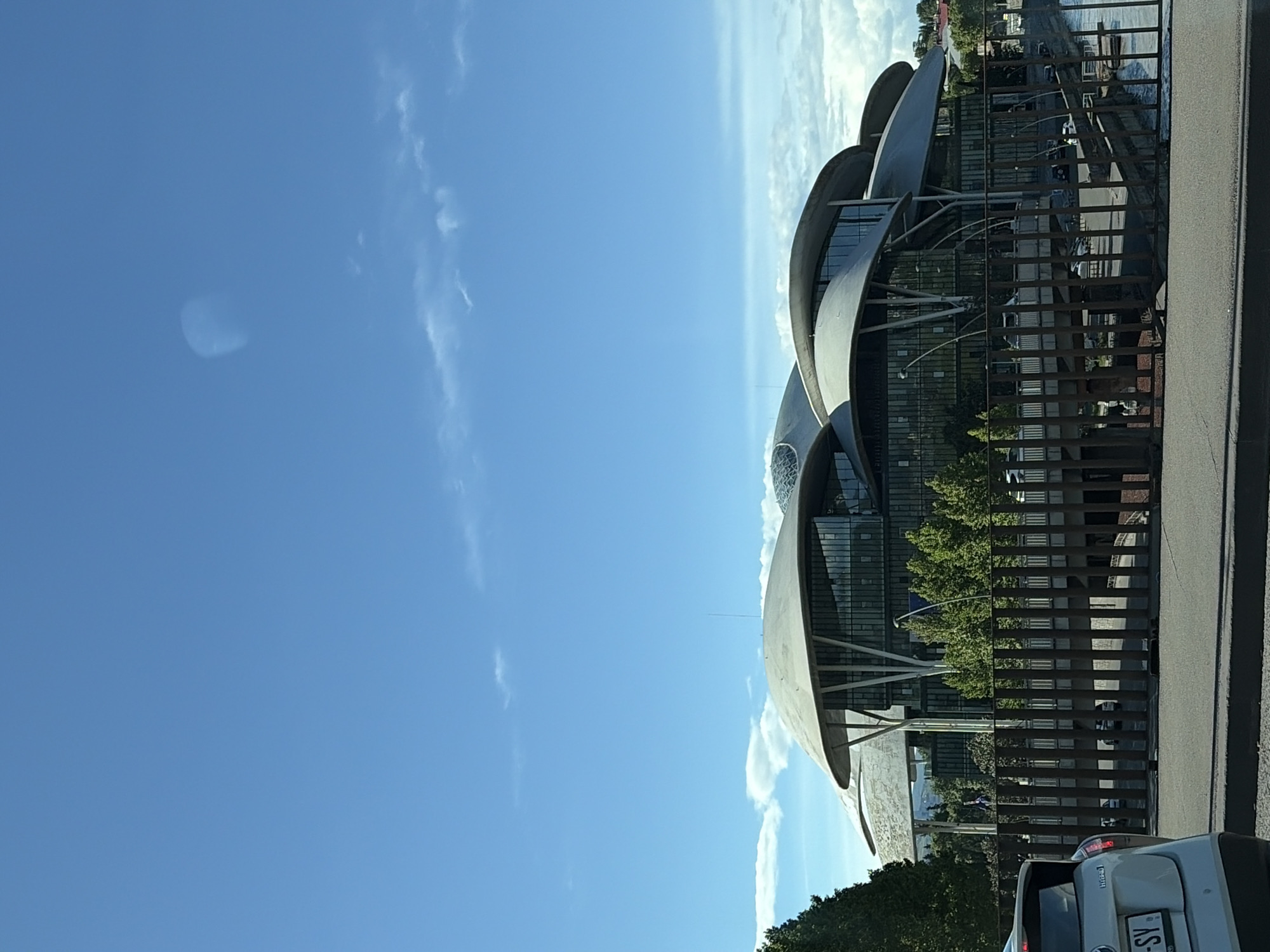 The image shows the Public Service Hall in Tbilisi, Georgia. The building has a distinctive architectural style with multiple overlapping, curved roof structures. The scene includes a partly visible car, a fence, and trees. The sky is blue with scattered clouds. The building appears to be located near a roadway.