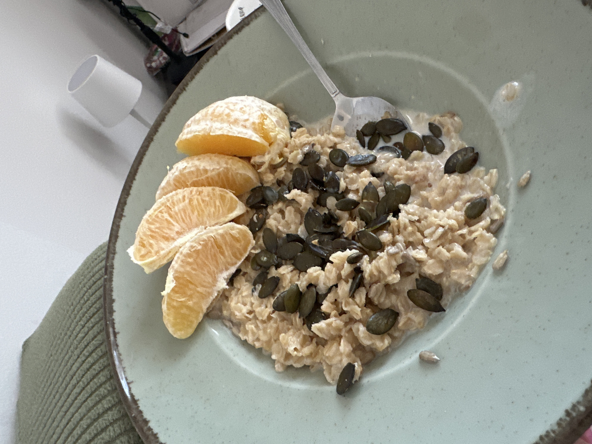 The image depicts a light green plate containing a breakfast dish. The dish consists of oatmeal, topped with dark green pumpkin seeds, and accompanied by sliced orange segments on the side. A silver spoon rests on the oatmeal. The plate is placed on a surface covered with a green knitted material. In the background, a white cylindrical lamp is visible, along with a stack of papers. The image was likely taken indoors, potentially in a residential setting, though the exact location in Novi Sad, Serbia, cannot be determined from the image alone. No people are directly visible in the image.