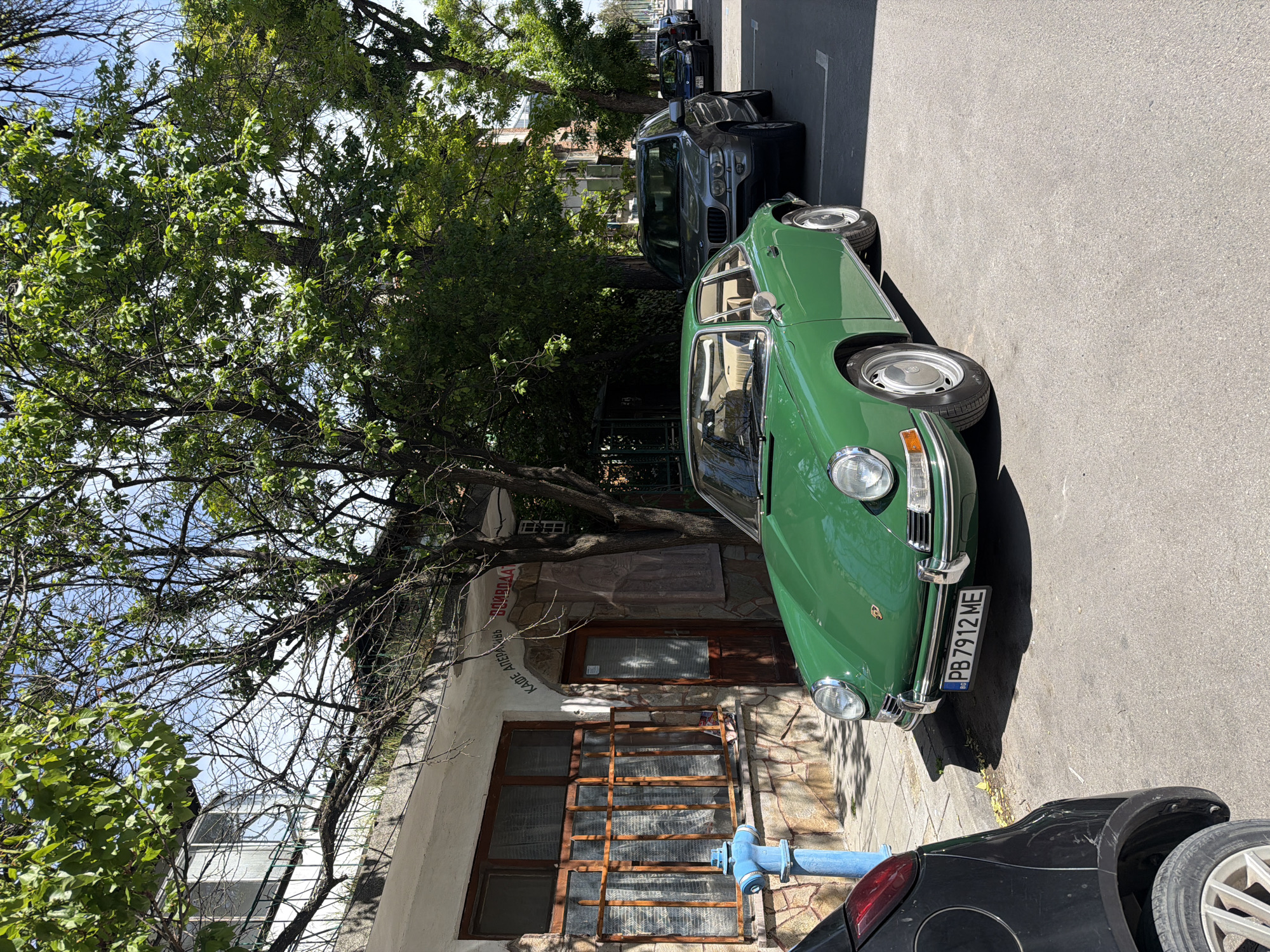 The image depicts a sunny street scene in an unidentified city. A green classic car, possibly a Porsche, is parked on the right side of the street, partially obscuring a black car in the foreground. Behind the green car are several other parked vehicles, including an SUV. To the left of the cars is a building constructed with a combination of stone and white stucco, featuring windows with metal grilles and a wooden door. A blue fire hydrant stands near the building. A large tree with dense foliage partially obscures the building and casts shadows onto the street. The license plate of the green car is visible and reads "PB 7912 ME".