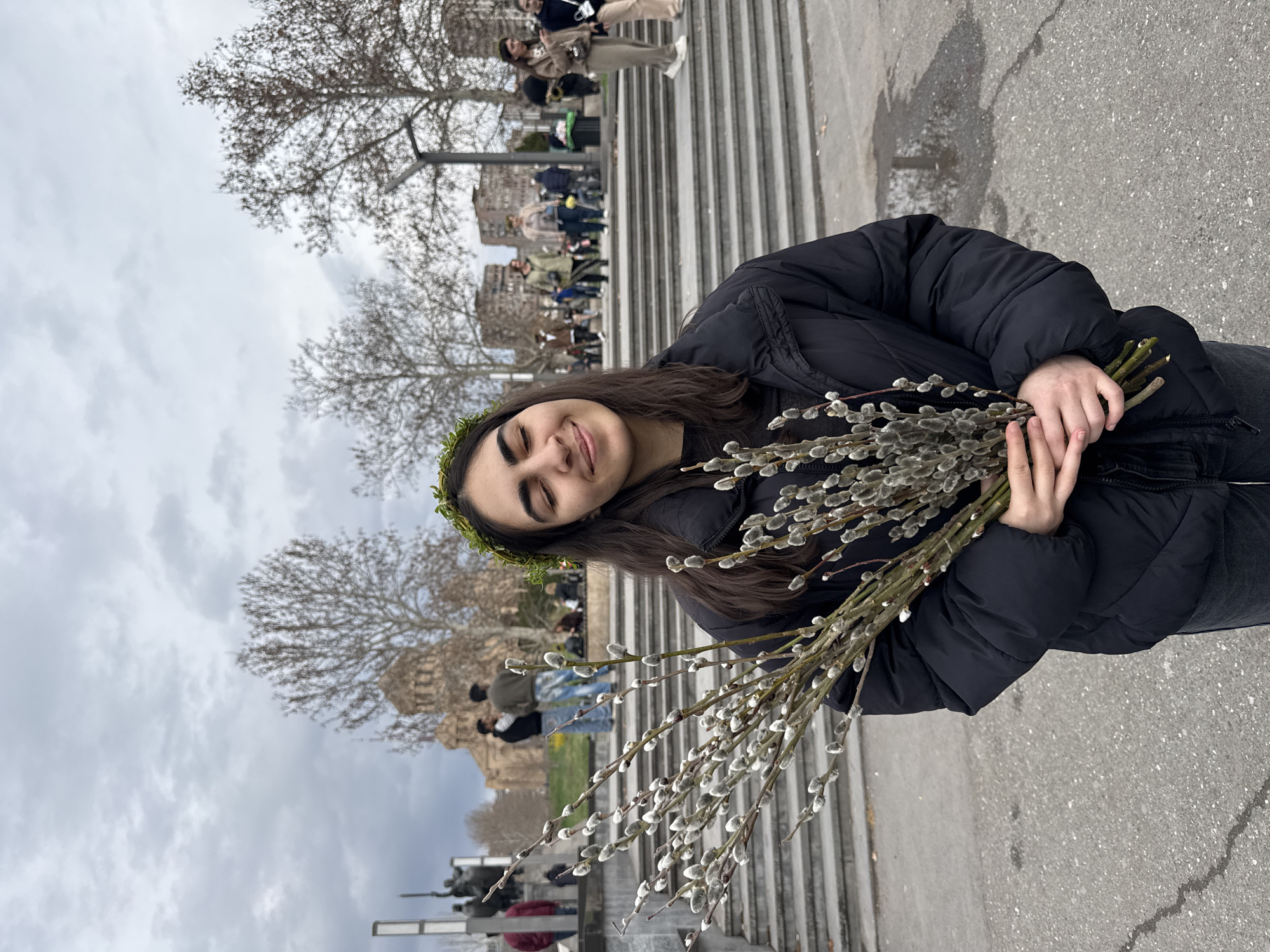 A young woman is holding a bunch of willow branches with soft buds, potentially pussy willows. She is smiling slightly with her eyes closed, suggesting contentment. She has a leafy crown on her head. She is wearing a black puffer jacket.
The photo appears to be taken outdoors, likely in a public space. In the background, there's a large staircase, and people are walking on it. A building is visible behind the people. The sky is overcast.
The background suggests the Cascade complex in Yerevan, Armenia.