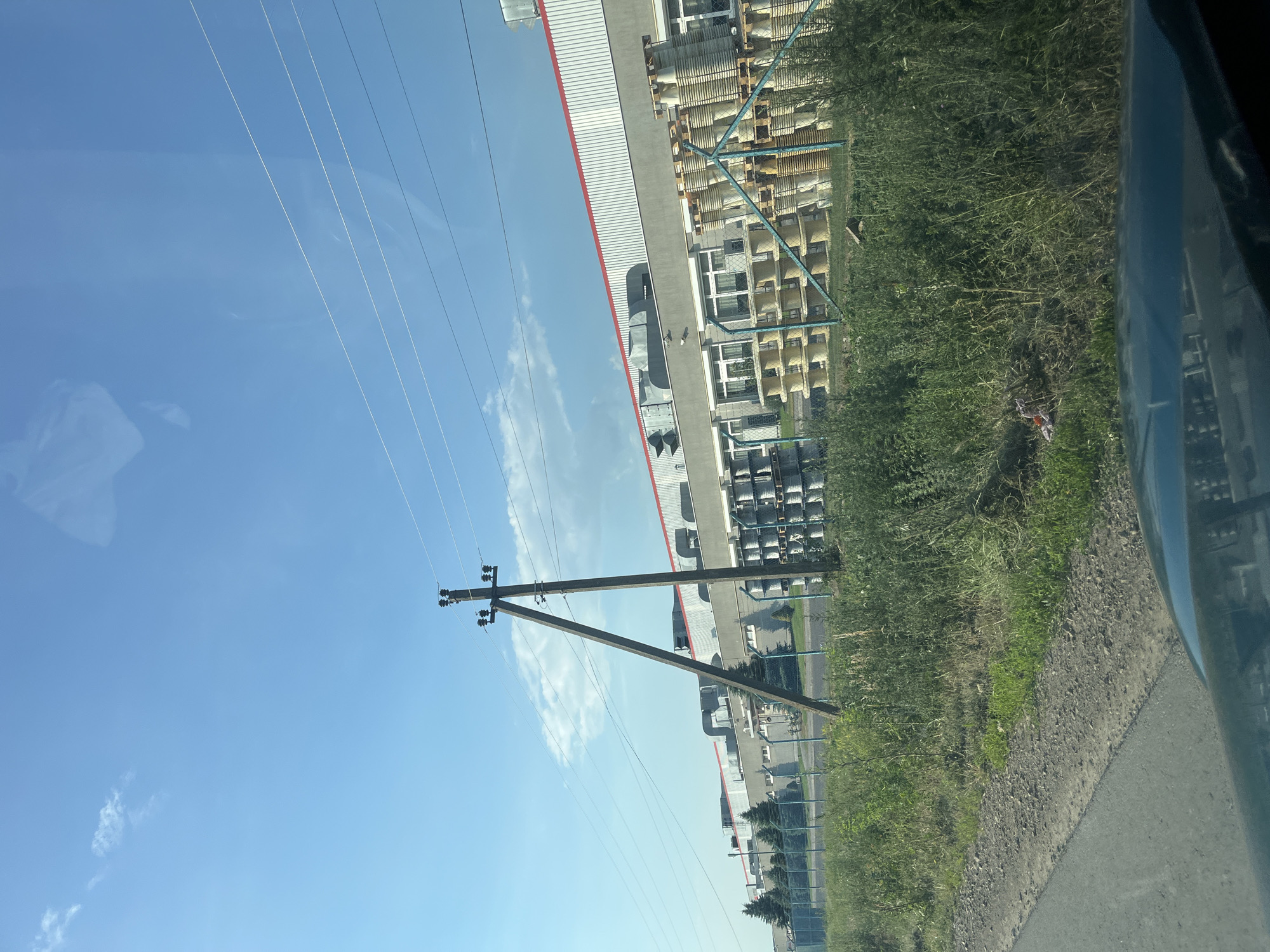 The image depicts an industrial area under a clear blue sky with scattered clouds. In the foreground, a dark-colored vehicle's hood is visible. To the right, a gravel road with overgrown grass lines its edge. A prominent wooden utility pole with electrical insulators stands in the middle ground. Behind the pole, a long, modern industrial building with a red-trimmed roof and grey walls is visible. The building features several large air conditioning units on its roof. In front of the building, behind a blue chain-link fence, numerous large, stacked spools of wire or cable are arranged, some on pallets. Several evergreen trees are planted in a line between the fence and the building. The overall scene suggests a manufacturing facility located in an industrial zone.