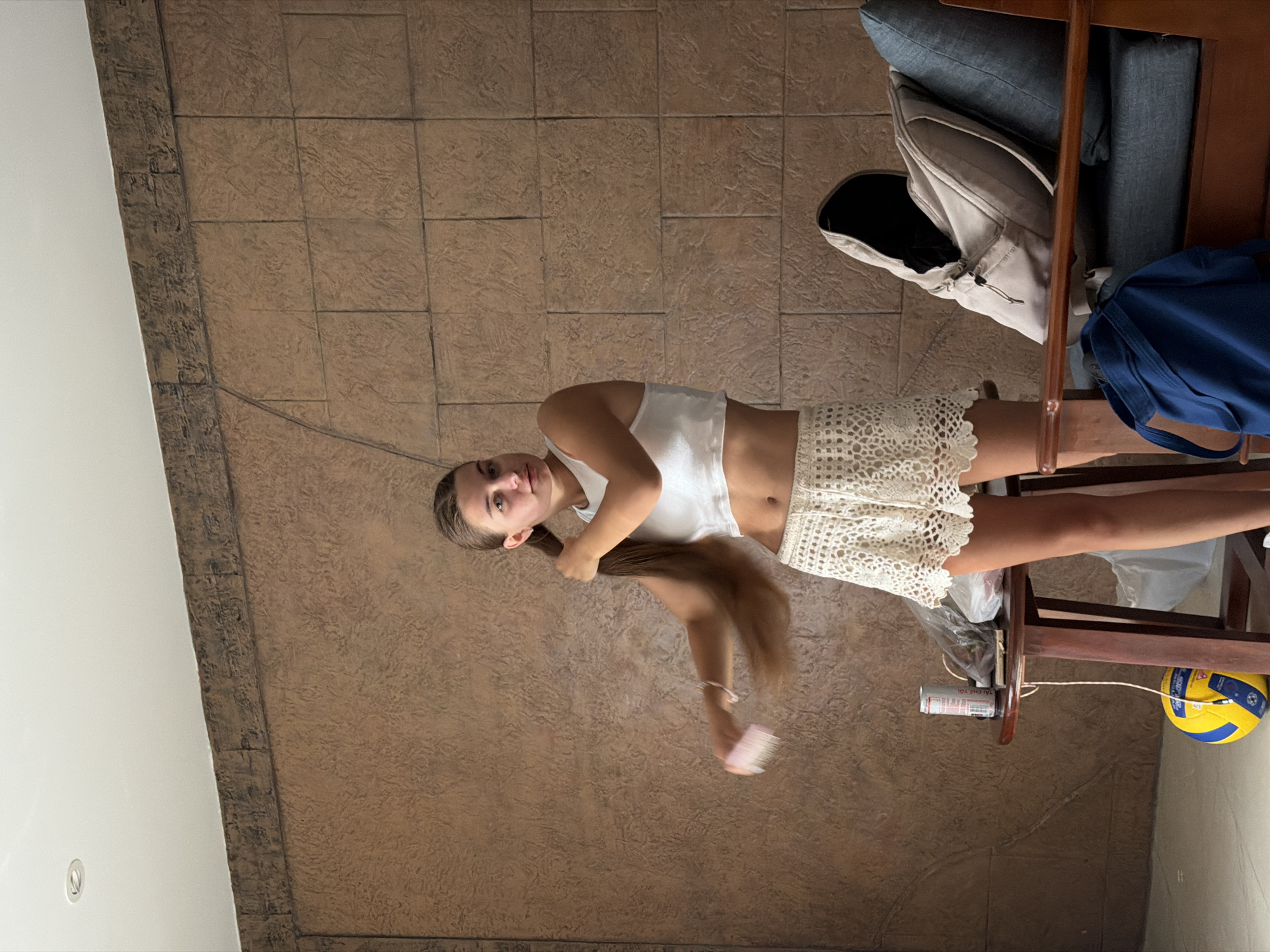 A young woman stands in a room with a textured brown wall and a white ceiling. She is wearing a white crop top and a white lace skirt. Her long brown hair is partially brushed with a pink comb. To her right, a wooden table holds a can, a bag of items, and a volleyball on the floor. A chair with blue cushions is to the right of the table, with a light-colored backpack on the seat and a blue bag on the floor in front of it. The scene appears to be indoors, possibly in a hotel or residence.