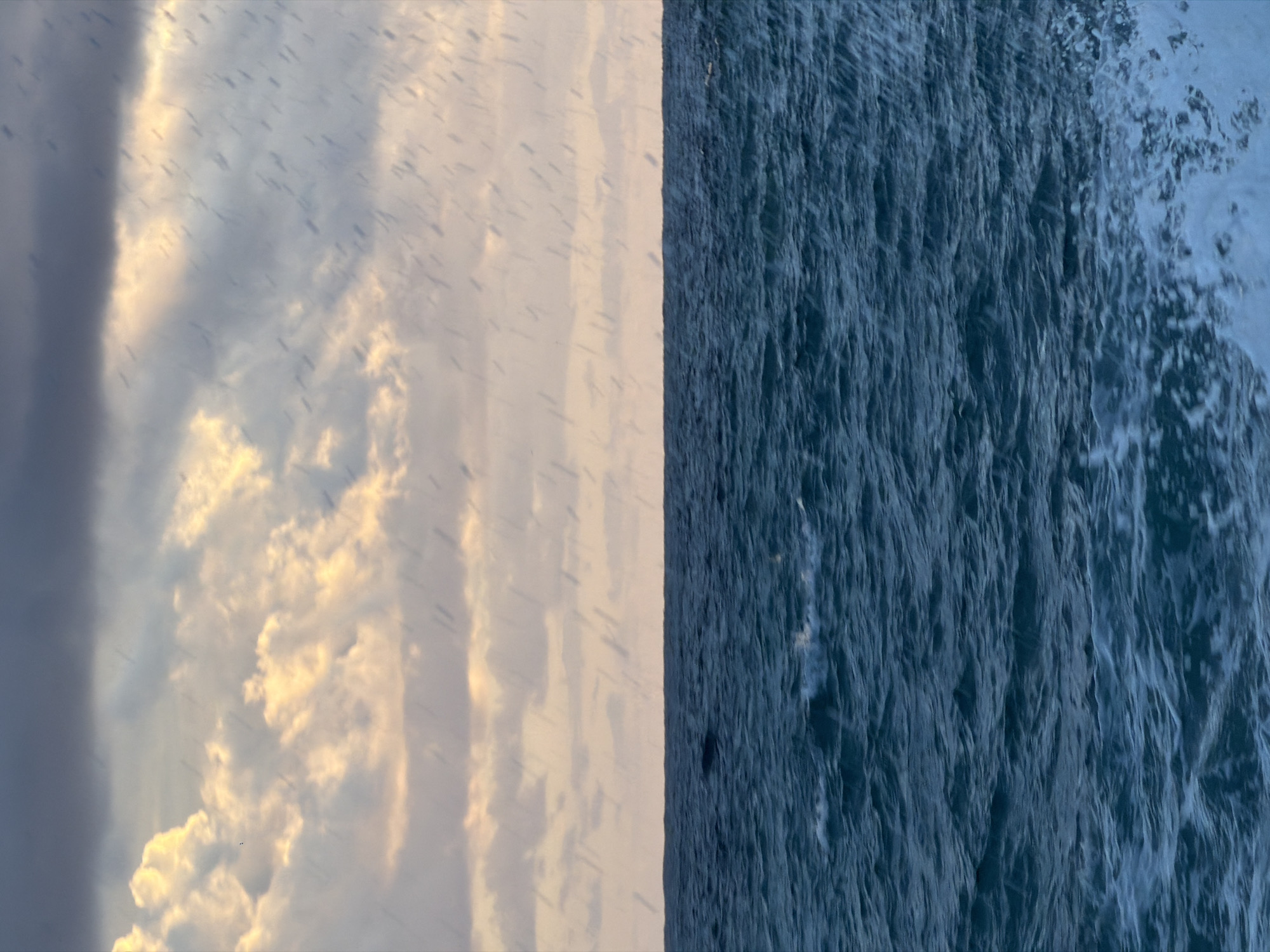 The image is vertically split into two distinct sections. The left side depicts a sky with varying cloud formations, ranging from dark gray and heavy to light, fluffy white clouds touched with gold tones. Dark streaks or patterns are visible across the sky. The right side displays turbulent, dark blue ocean water with white foam visible in the top right corner. The overall impression is one of juxtaposed weather conditions, where a stormy sky meets a rough sea. There is no clear evidence in the image to confirm that it is located in Petrovac centar, Montenegro. Therefore, I can not provide any location information.