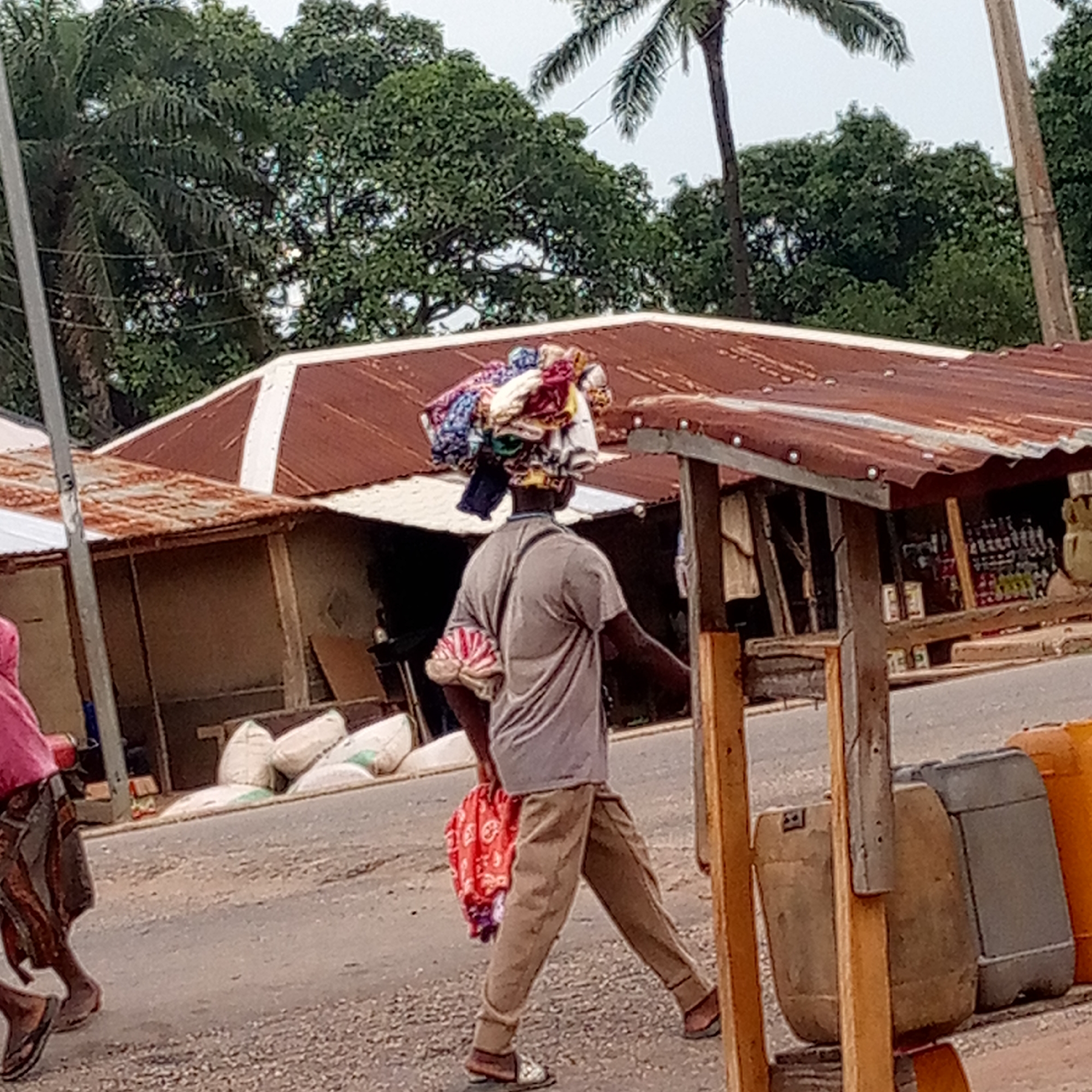 A person is walking on a paved road in Mararaba, Nigeria. The person is carrying a stack of fabric on their head and another bundle in their left hand. The person is wearing a light gray shirt, tan pants, and sandals. In the background, there are various buildings with corrugated metal roofs. There are also trees and vegetation in the background, including a palm tree. To the right, a wooden structure with a corrugated metal roof houses various items, including bottles and containers. To the left, another person wearing a pink shirt walks past. The road surface appears to be weathered. There are bags of goods in the background, resting near the side of the road by a building.
