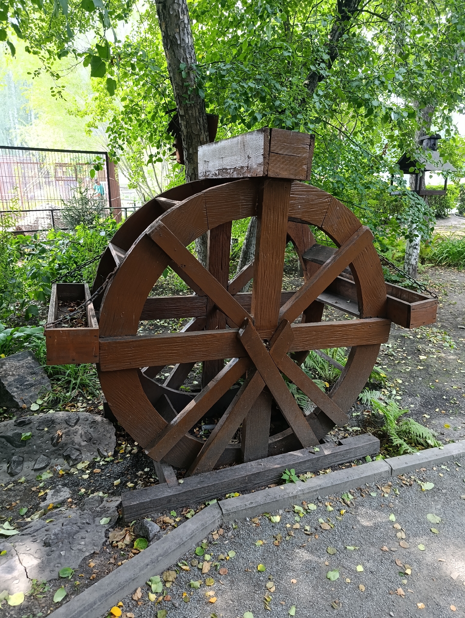A wooden water wheel, painted brown, stands prominently in a park setting. The wheel is large and features a spoke design. Attached to the sides of the wheel are wooden troughs, which appear to be empty. The water wheel is supported by wooden beams resting on the ground. The surrounding environment is lush with green foliage from trees and bushes. Fallen leaves are scattered on the ground, which is covered with gravel and features a concrete curb. In the background, a fence and a bird feeder are visible among the trees. The scene appears to be a decorative element in a public outdoor space.
