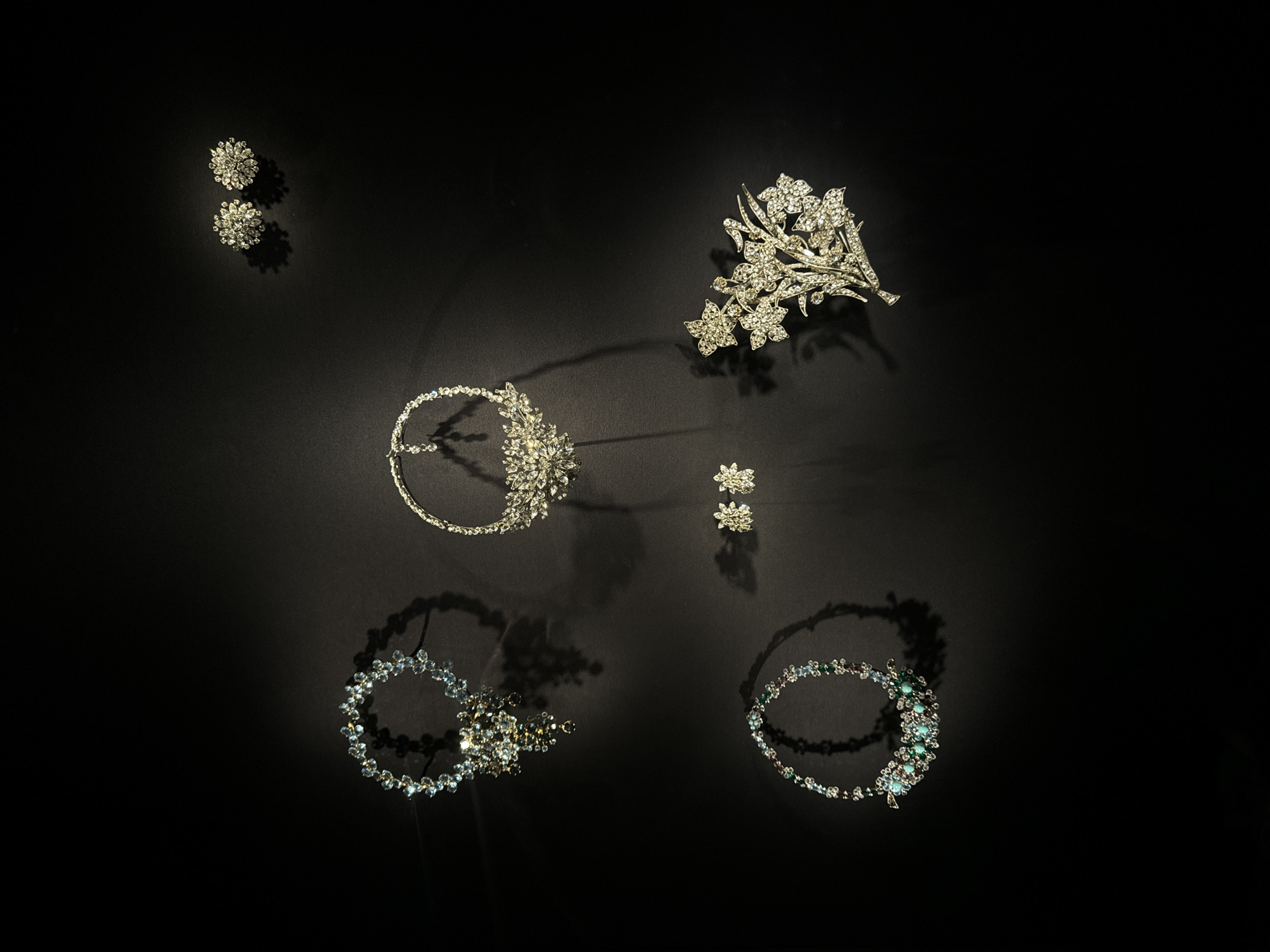 A curated collection of sparkling jewelry is arranged on a deep black surface, bathed in focused, warm lighting. The assortment includes floral earrings, a jeweled headband, a flower-shaped brooch, and a pair of delicate gemstone bracelets featuring hints of turquoise and green. These bracelets appear to be laid in a circular pattern.
The image's setting is unknown and there is no location information present. It is not possible to ascertain if the photo was taken in Paris, France.