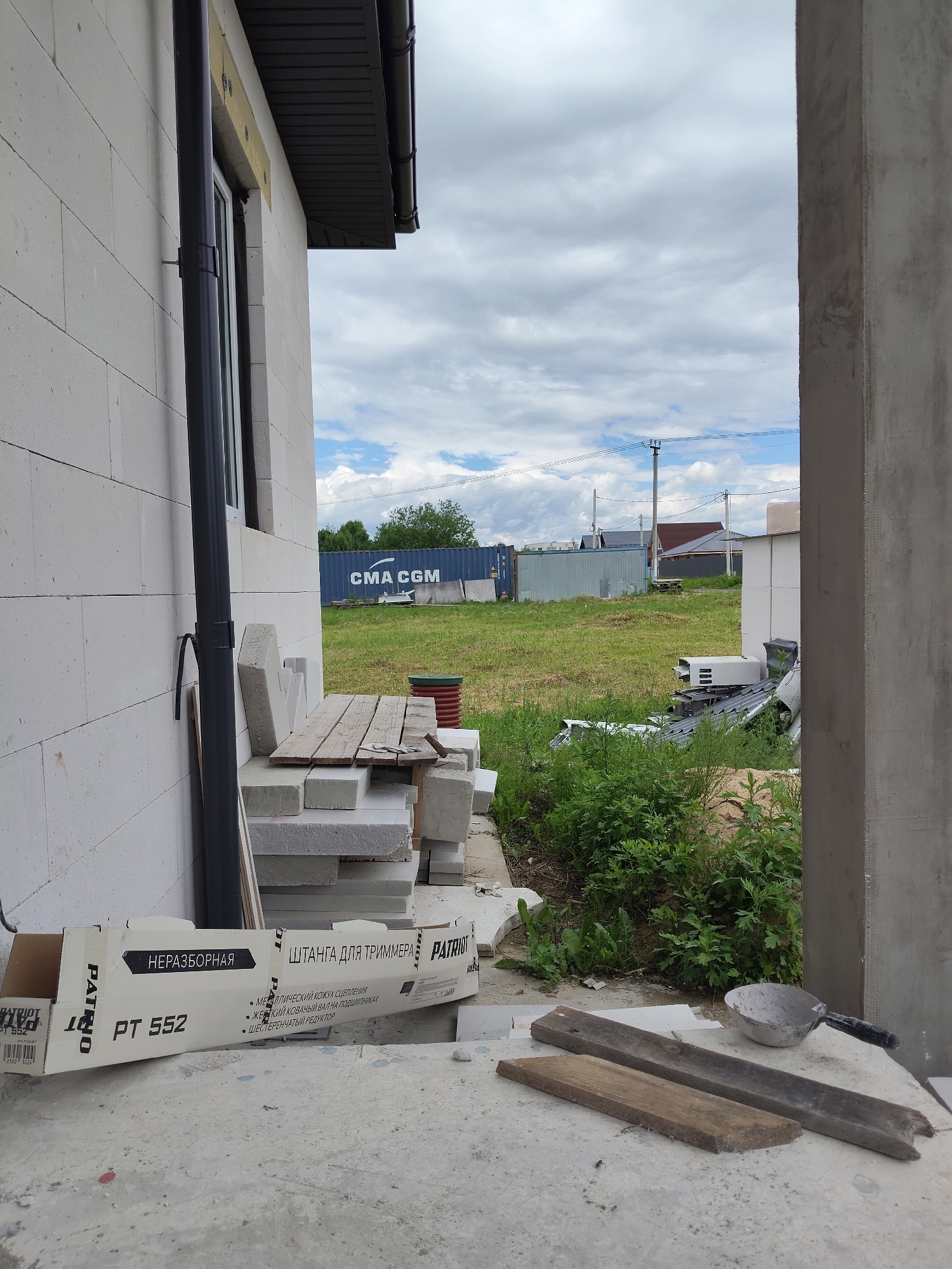In Skurygino, Russia, an exterior shot captures a construction site on a cloudy day. A pale brick building, possibly under construction, dominates the left side of the frame, featuring a dark downspout running vertically. In the foreground, a cardboard box with Russian text, including "ПАТРИОТ" and "PT 552," lies on a concrete surface. To the right, a concrete pillar marks the edge of a building or structure. Beyond the immediate construction area, a grassy field is visible, dotted with green vegetation. In the middle ground, a blue shipping container with "CMA CGM" printed on it stands alongside a light gray fence. Further in the distance, houses with dark roofs and utility poles with power lines suggest a residential or semi-rural area. Several stacks of concrete blocks and wooden planks are also present on the site, indicating ongoing construction work.