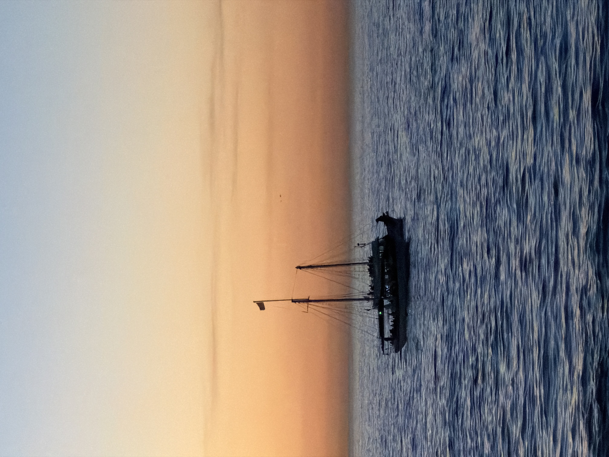 The image depicts a sailboat on the ocean at sunset. The sky transitions from light blue to orange. The sailboat is dark, casting a faint shadow on the water. The ocean has many distinct dark blue wave peaks. No people can be identified. The location is not explicitly evident as Cape Town, South Africa.