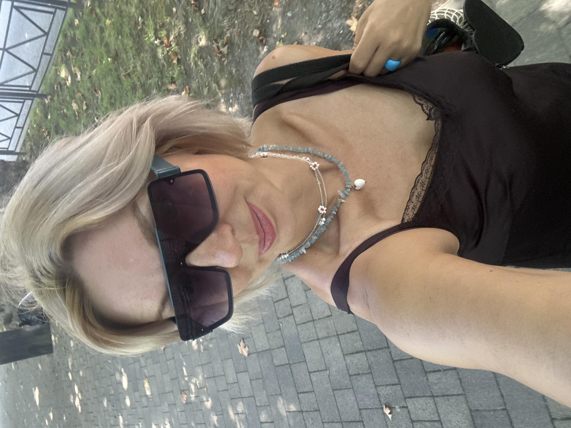 A blonde woman wearing large, dark sunglasses and a dark camisole with lace trim takes a selfie outdoors. She has on a layered necklace with light blue beads, white flowers with red centers, and a white heart pendant. A bright blue ring is visible on her right hand. She is carrying a black bag with a heart-shaped design. The background shows a paved walkway, green grass, and a black metal fence. The overall lighting suggests it is daytime. The environment, characterized by the paved walkway and the style of the fence, is consistent with a park or promenade area within the city of Sochi, Russia.