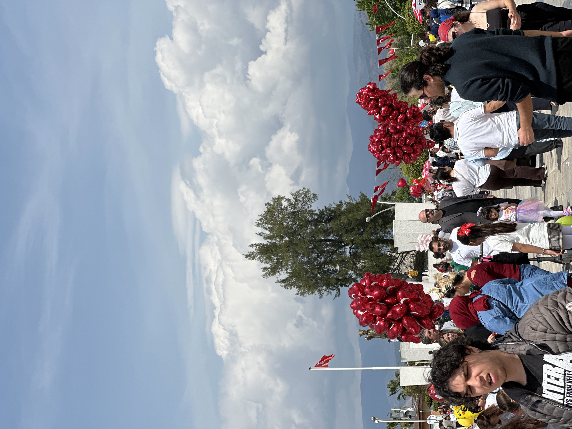 The image depicts an outdoor public gathering in Fethiye, Turkey, under a bright, partly cloudy daytime sky. A diverse crowd of people, including adults and children, fills the scene, primarily observing an unseen event or activity to the right of the frame. The atmosphere is festive and patriotic, highlighted by numerous large bunches of red heart-shaped balloons, many of which bear the white crescent moon and star symbol of the Turkish flag.
Small Turkish flag pennants are strung across the area, and a prominent Turkish flag flies from a flagpole in the mid-ground. In the foreground, a young man with dark, curly hair is visible, wearing a black t-shirt with the partial text "YS FROM HELL". Other individuals in the crowd include a man wearing sunglasses and a suit, and a small girl in a colorful tutu. Behind the gathering, green trees and parts of a light-colored building are visible, with distant mountains forming a scenic backdrop under the clear, sunny sky with large white clouds. The setting suggests a public square or promenade, indicative of a national holiday or celebratory event.
