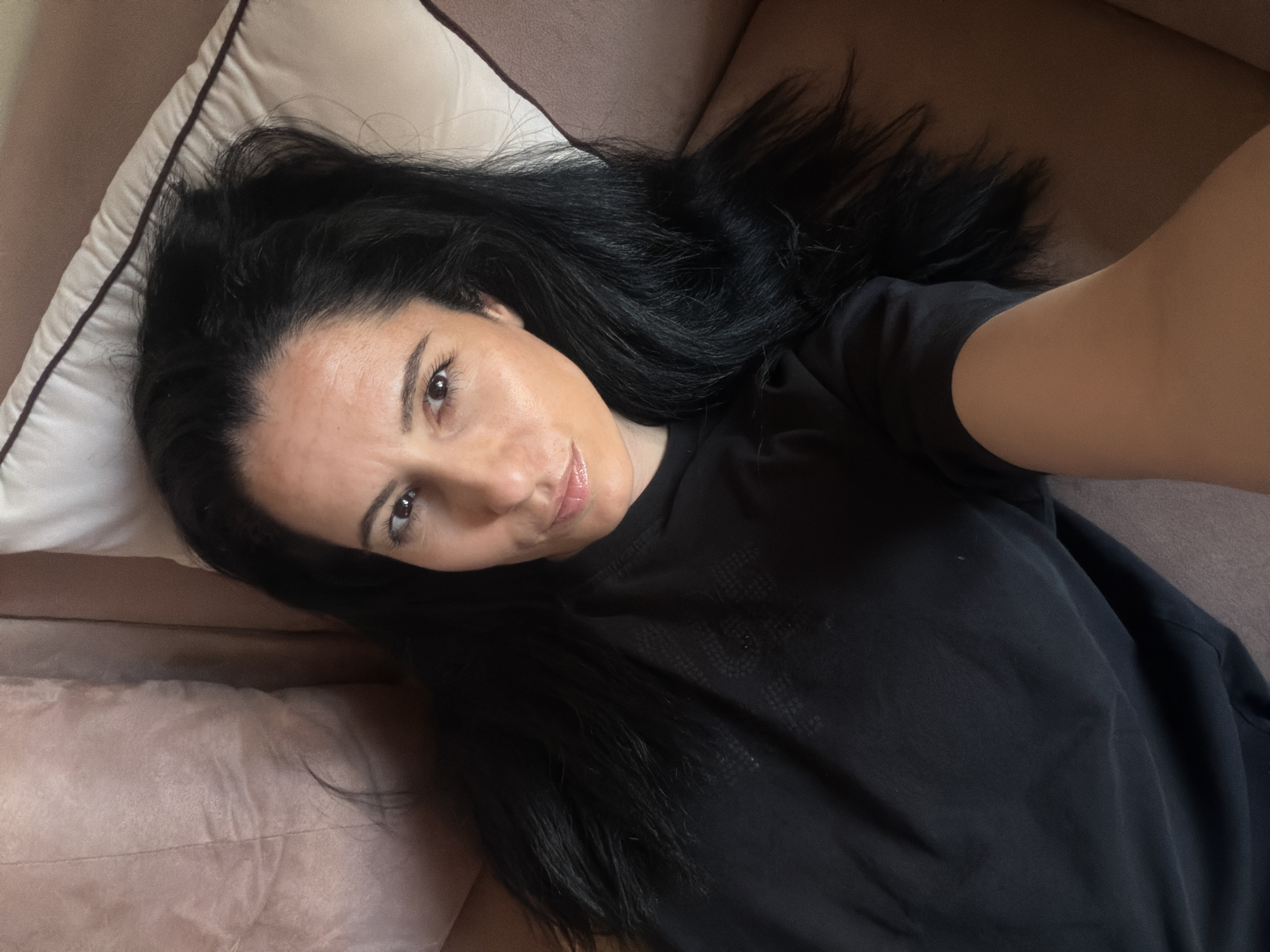 A woman with long, dark hair is lying on a pink couch with a white pillow. She is wearing a black shirt and looking at the camera, taking a selfie. There is no indication of the location in Pleven, Bulgaria.