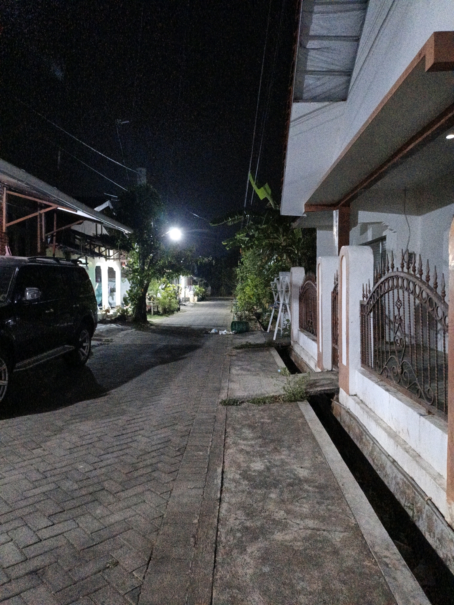 A nighttime street view in Cikal, Indonesia shows a parked black SUV on the left, positioned on a brick-paved driveway. The driveway extends into the distance, leading past houses and trees illuminated by a streetlamp. On the right, a white house features a decorative iron fence and a concrete walkway bordering a drainage ditch. The scene is generally dark, with ambient light coming from the streetlamp and interior lights of the houses.