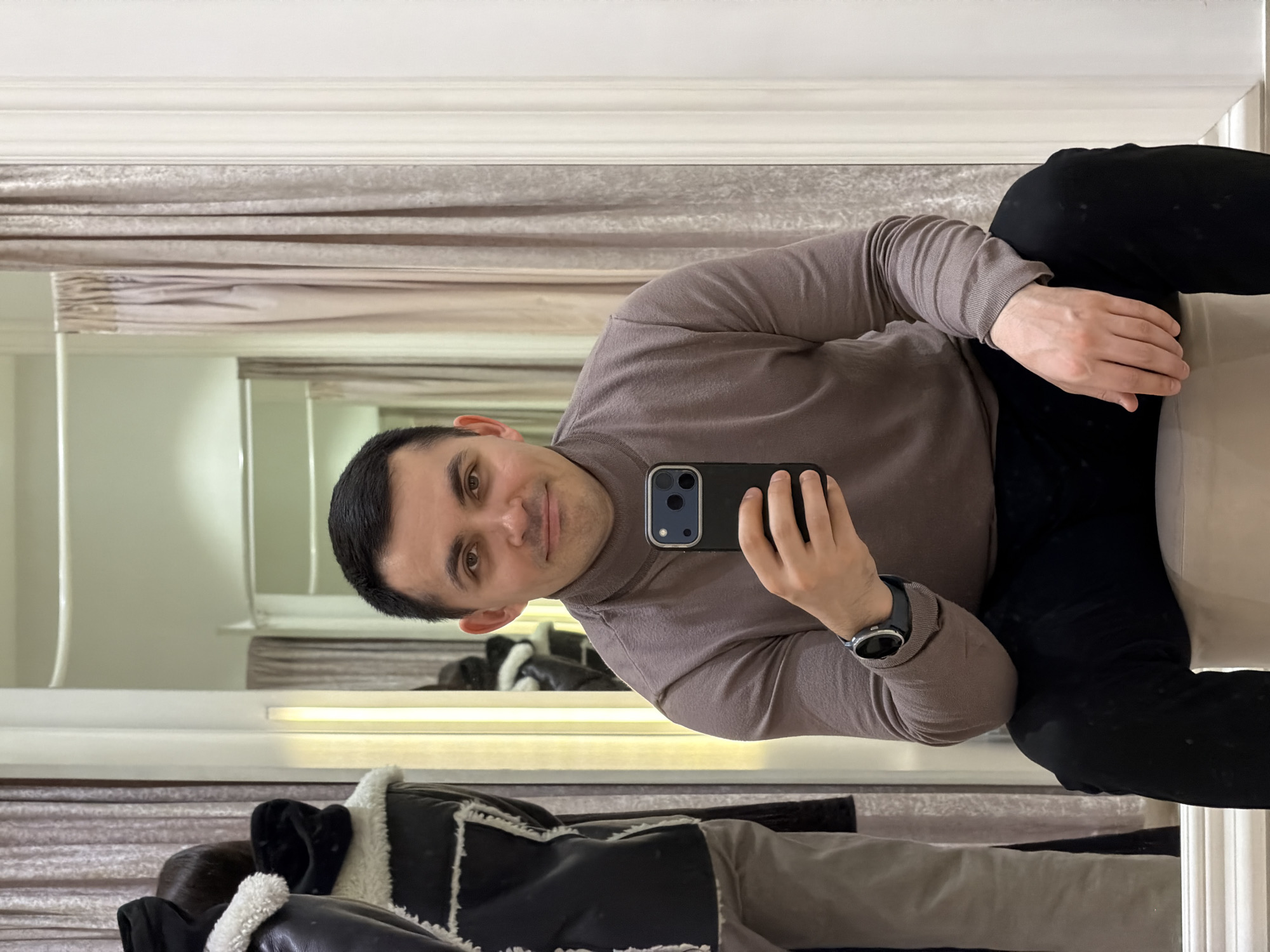 A person is taking a selfie in a mirror. The person is wearing a brown turtleneck sweater, black pants, and a watch. The person is holding a black smartphone in their right hand. The mirror is framed by a white wooden frame. The reflection in the mirror shows a white wall, a beige curtain, and a partially visible jacket. The location cannot be identified as Izhevsk, Russia, based on the visual information provided.