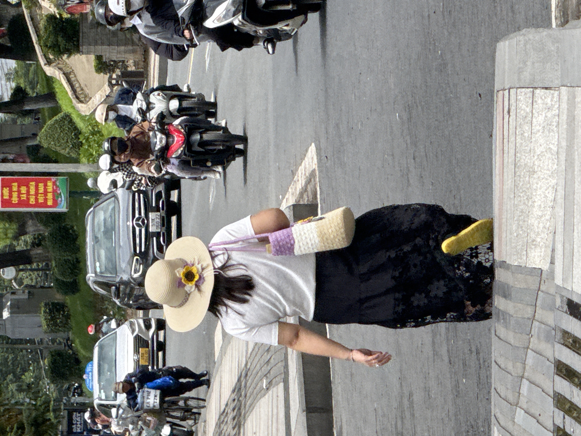 This image captures a bustling street scene in Da Lat, Vietnam, likely on a main thoroughfare. The foreground is dominated by a woman from the back, wearing a wide-brimmed straw hat adorned with a sunflower and purple accents, a white t-shirt, and a long black skirt with floral lace details. She is carrying a woven purple, white, and beige bag and is wearing bright yellow shoes. She appears to be walking away from the viewer, ascending a set of concrete steps that are part of the city's infrastructure.
The middle ground features a dynamic flow of traffic. Several motorbikes are visible, with riders wearing helmets and masks, common in this region. One motorbike in red is prominent. Behind the motorbikes, a silver car and a dark SUV are on the road. A man is seen pushing a bicycle loaded with items, including what appears to be bags labeled "CA PHE" (coffee).
The background shows a vibrant urban environment with green trees and lush foliage lining the street, characteristic of Da Lat's hilly terrain. A red and white billboard with Vietnamese text is also visible on the left side of the image. The overall impression is one of daily life and movement in a Vietnamese city.