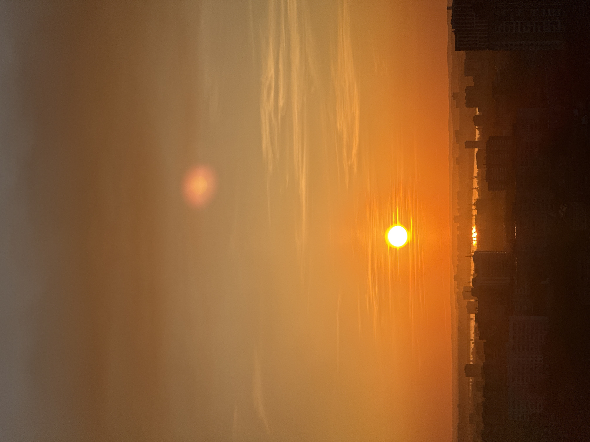 The image depicts a cityscape at sunrise or sunset. The sun is a bright, hazy orb positioned in the center of the frame. The sky is filled with orange and yellow hues, suggesting either sunrise or sunset, possibly due to atmospheric conditions like pollution or dust. Silhouettes of buildings and structures are visible along the horizon on the right, suggesting an urban environment. It appears to be a view overlooking the city from a high vantage point. There is no evidence to confirm this location is in Perm, Russia.