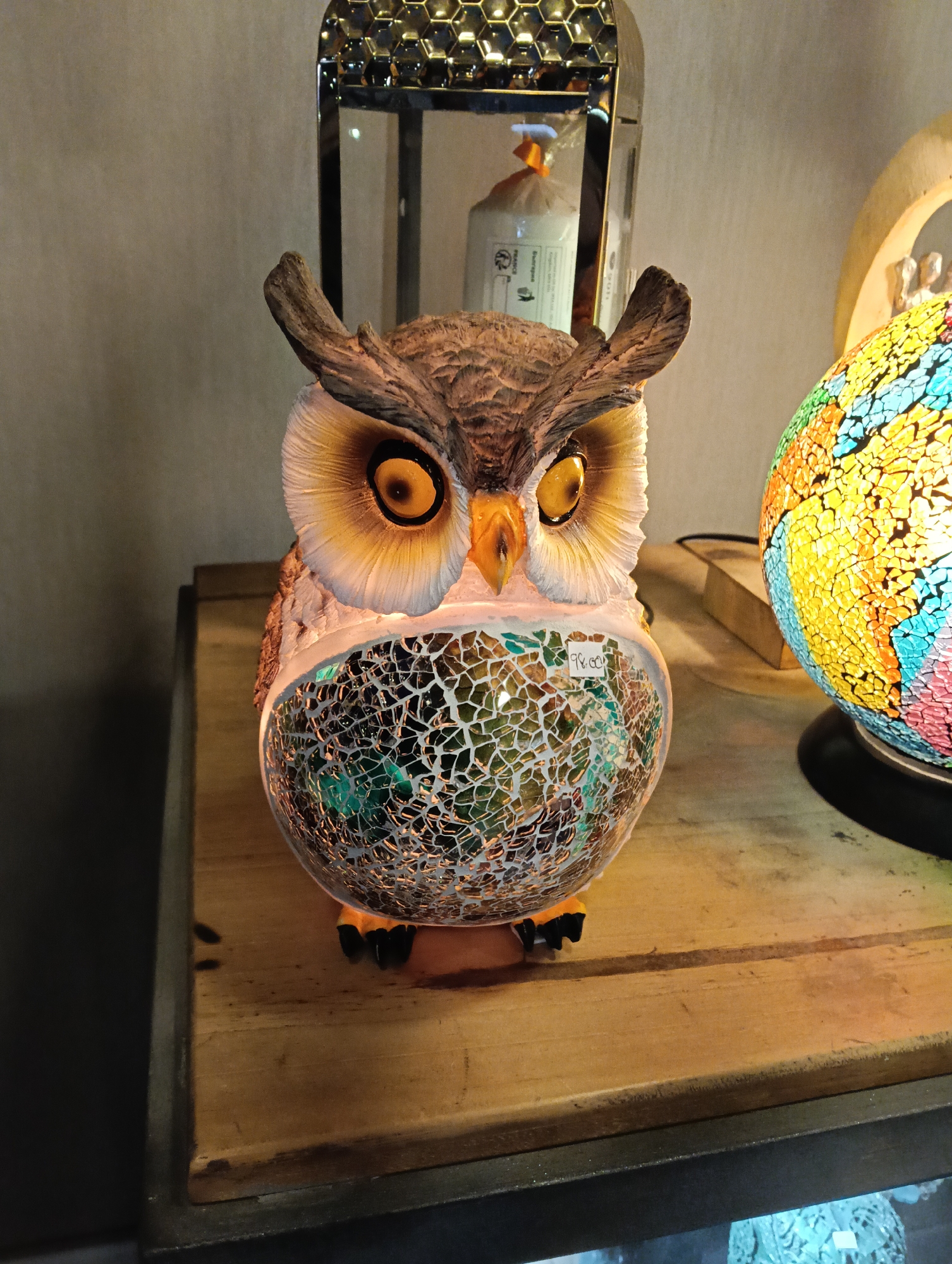 An indoor shot features a decorative owl lamp as the central subject, resting on a wooden surface. The owl has an intricately detailed head with brown and white feathers, prominent yellow eyes, and a small beak. Its body is composed of a translucent mosaic of cracked glass in shades of green, white, and brown, illuminated from within, creating a warm glow. A small white price tag sticker reading "98.00" is affixed to the owl's mosaic body.
To the right of the owl, a colorful spherical lamp is visible, made of numerous mosaic pieces in yellow, orange, blue, green, and red, also glowing from an internal light source. Behind the owl, a tall, metallic, hexagonal decorative object or lamp stands, with a honeycomb-like pattern on its top. Inside this object, a white, plastic-wrapped item with some indistinct text is visible.
The wooden surface supporting these items appears to be a shelf or table, showing some grain and wear. The lighting is artificial and warm, highlighting the illuminated decorative items and creating a cozy ambiance. The scene is likely set in a shop or home decor display in Puerto Del Carmen, Spain.