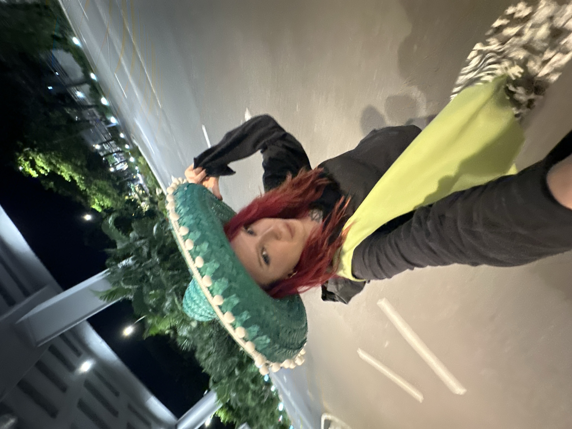 A woman with red hair and dark eye makeup is posing for a selfie outdoors at night, wearing a large, teal sombrero adorned with white pompoms. She is dressed in a black long-sleeved shirt, a bright yellow vest, and a patterned scarf. The background features a dimly lit urban environment with palm trees, streetlights, and a road with white lane markings. The scene appears to be near a covered walkway or overpass, suggesting a location in Đà Nẵng, Vietnam. The woman is holding the brim of her sombrero with her right hand, and her left arm is bent. The lighting suggests it is nighttime or late evening.