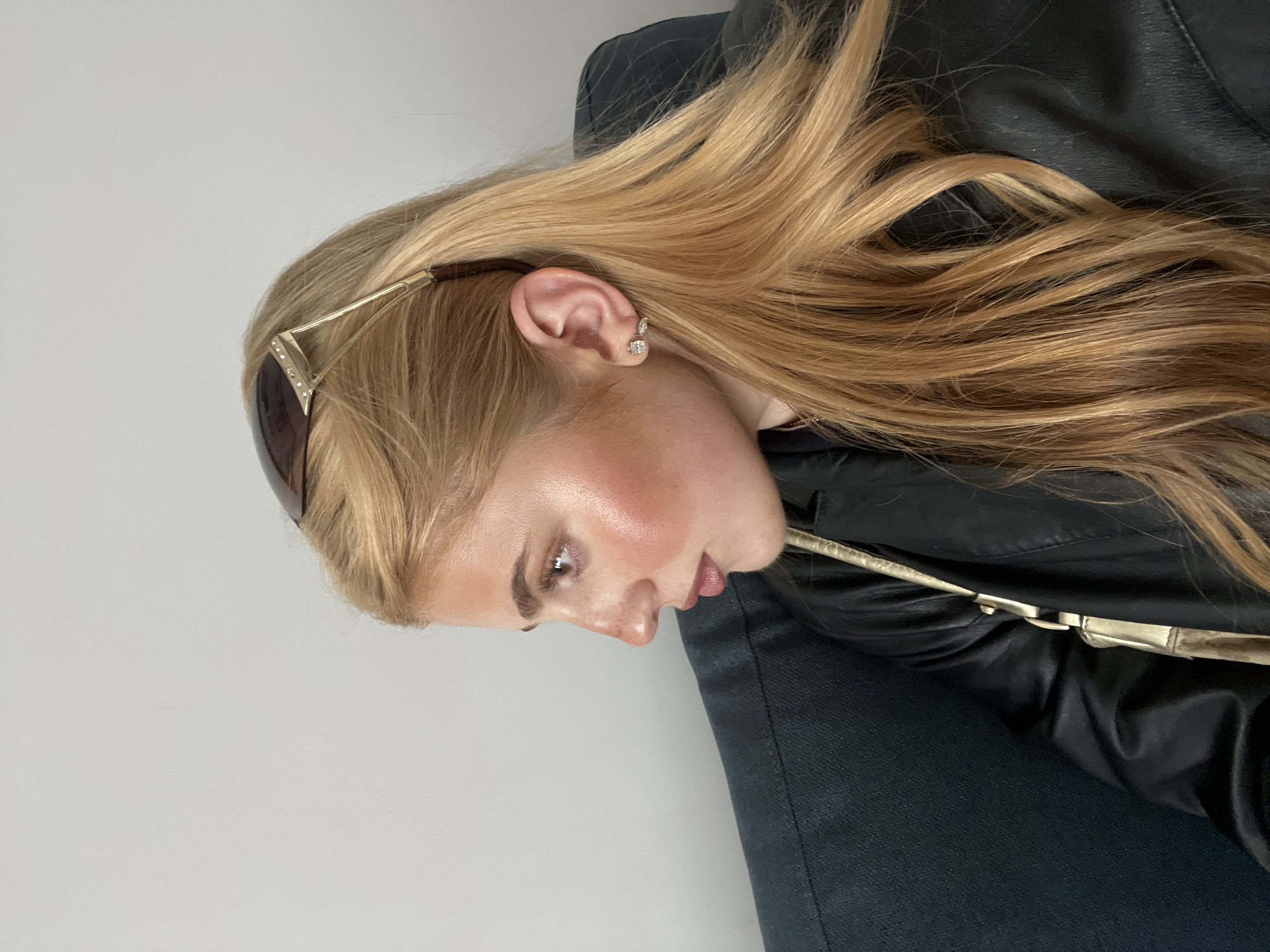 A fair-skinned female with long blonde hair is visible in a medium shot, partially reclined on a dark-colored fabric surface that appears to be a couch or seat. Her head is turned to the side in profile, looking downwards. She is wearing sunglasses pushed back on her head and earrings on her left ear. She is wearing a black leather jacket, with a gold-colored strap visible over her shoulder. The background is a plain, light gray or white surface. There is no direct indication of a location in Paris, France.