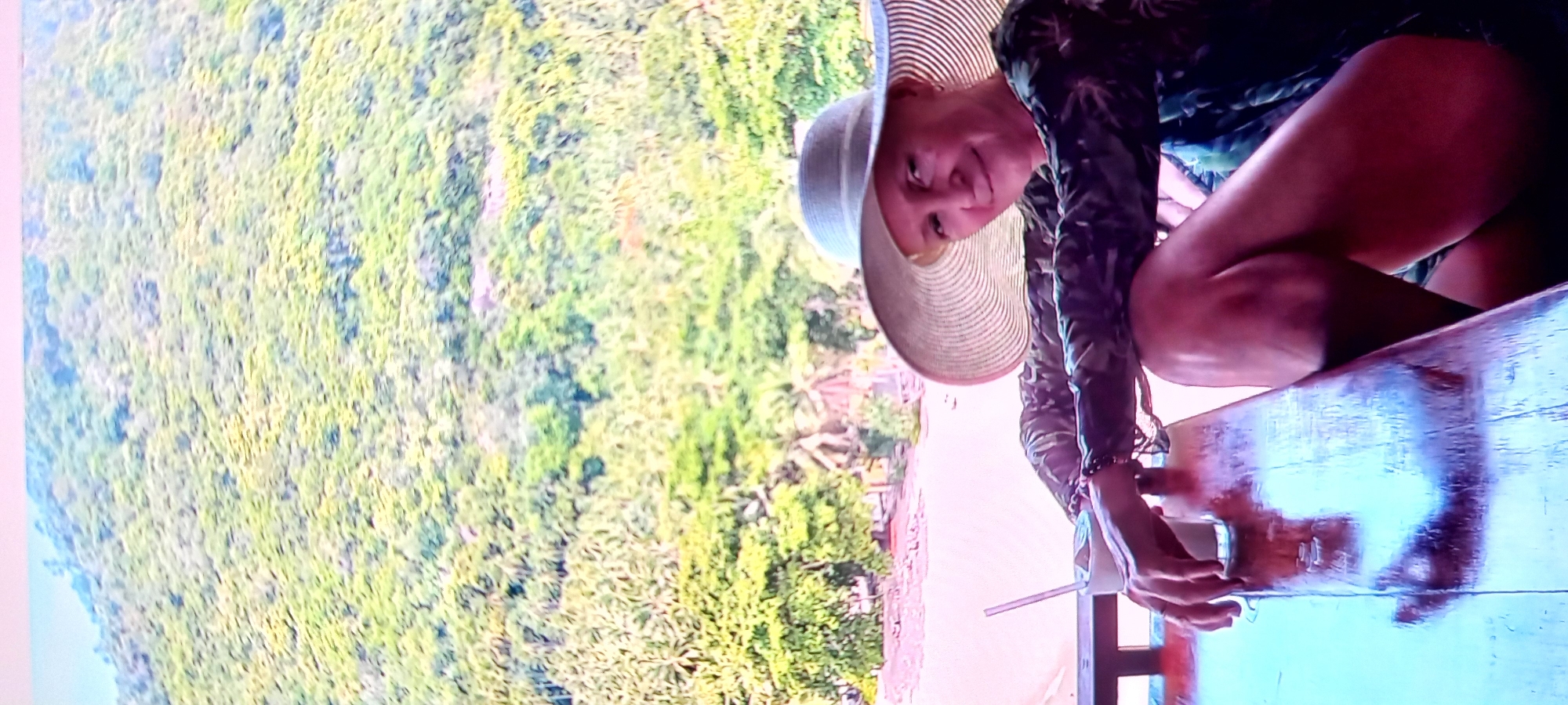 The image shows a woman in Baan Chaloklum, Thailand. She is wearing a wide-brimmed hat and a dark, patterned top. She is seated at an outdoor table, possibly on a balcony or deck, overlooking a lush, green hillside and a beach. The lighting suggests it is daytime. The view suggests a tropical or subtropical environment. The woman is holding a disposable cup and straw on the table.