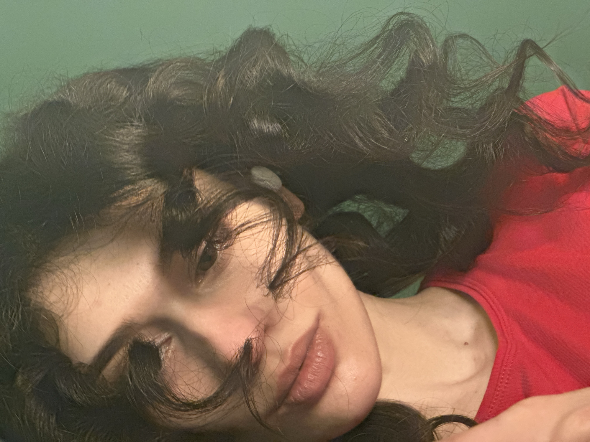 A close-up, low-angle shot shows the face and upper chest of a young woman with long, dark, curly hair. Her head is tilted to her left, and strands of hair fall across her face, partially obscuring her eyes. She is wearing a bright red top with a V-neck. Her skin is fair, and her lips are slightly parted. Her eyes are looking downwards, and the lighting casts shadows on her face, highlighting her cheekbones and the curve of her lips. The background is a solid, muted green. There is no indication of the image being taken in Sofia, Bulgaria, or any other specific city.