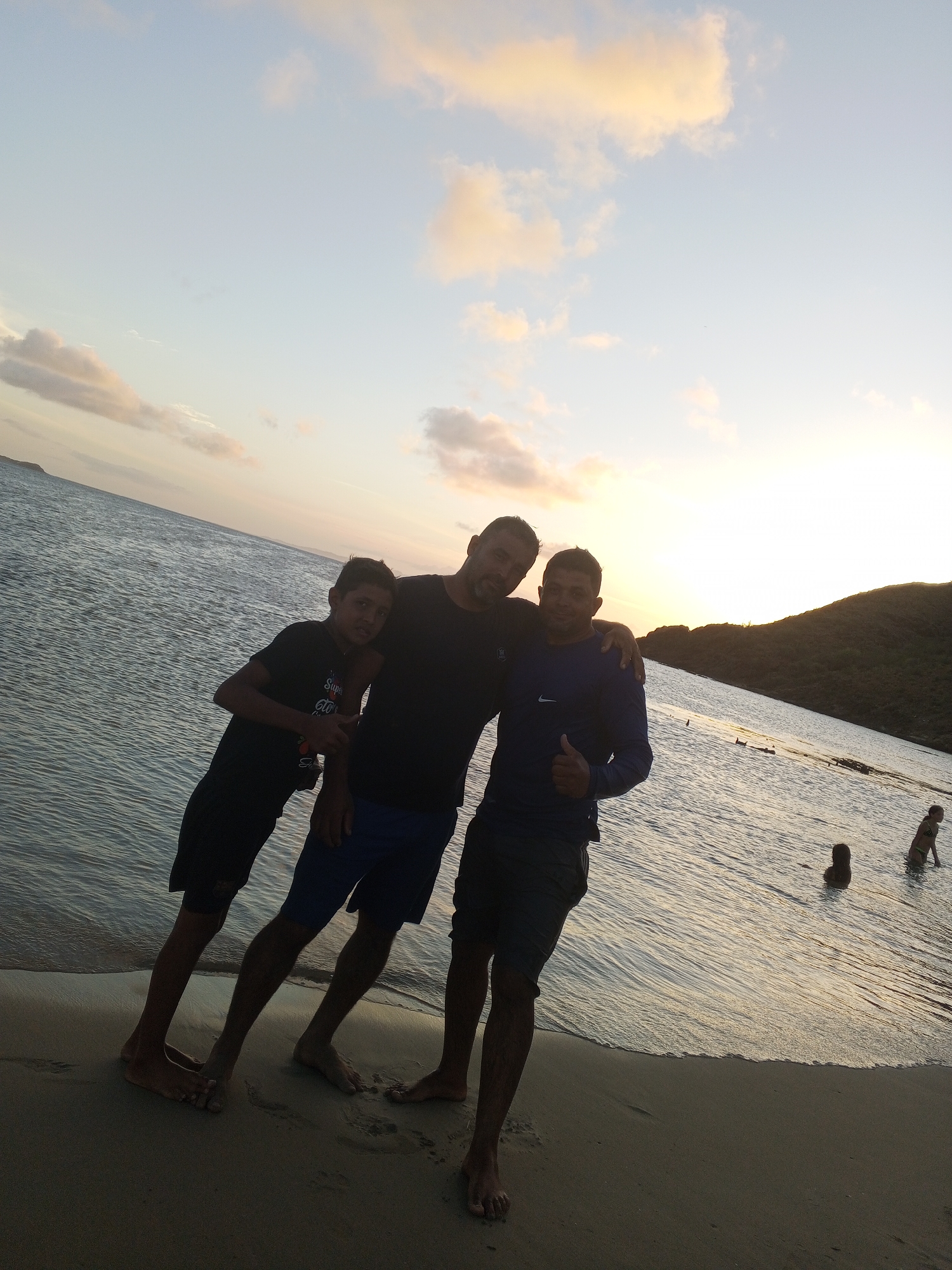 A group of three young men pose for a photo on a beach at Juan Griego, Venezuela. The man on the left, wearing a black t-shirt and black shorts, stands with his arm around the shoulders of the man in the center. The man in the center wears a black t-shirt and blue shorts, and has his arm around the man on the right. The man on the right wears a blue long-sleeved shirt and dark shorts, and gives a thumbs-up. The three men are standing at the edge of the water, with waves gently lapping at the shore.
In the background, two young girls are playing in the water, their figures silhouetted against the setting sun. The sky is a soft gradient of blue and pale yellow, with scattered clouds tinged with pink and orange, indicating that the photo was taken at sunset. The water is calm, reflecting the colors of the sky. A hilly, tree-covered landscape is visible in the distance, silhouetted against the bright horizon. The mood of the photo appears joyful and relaxed, with the friends enjoying a beautiful evening at the beach. The text "Super 600" is visible on the t-shirt of the man on the left.