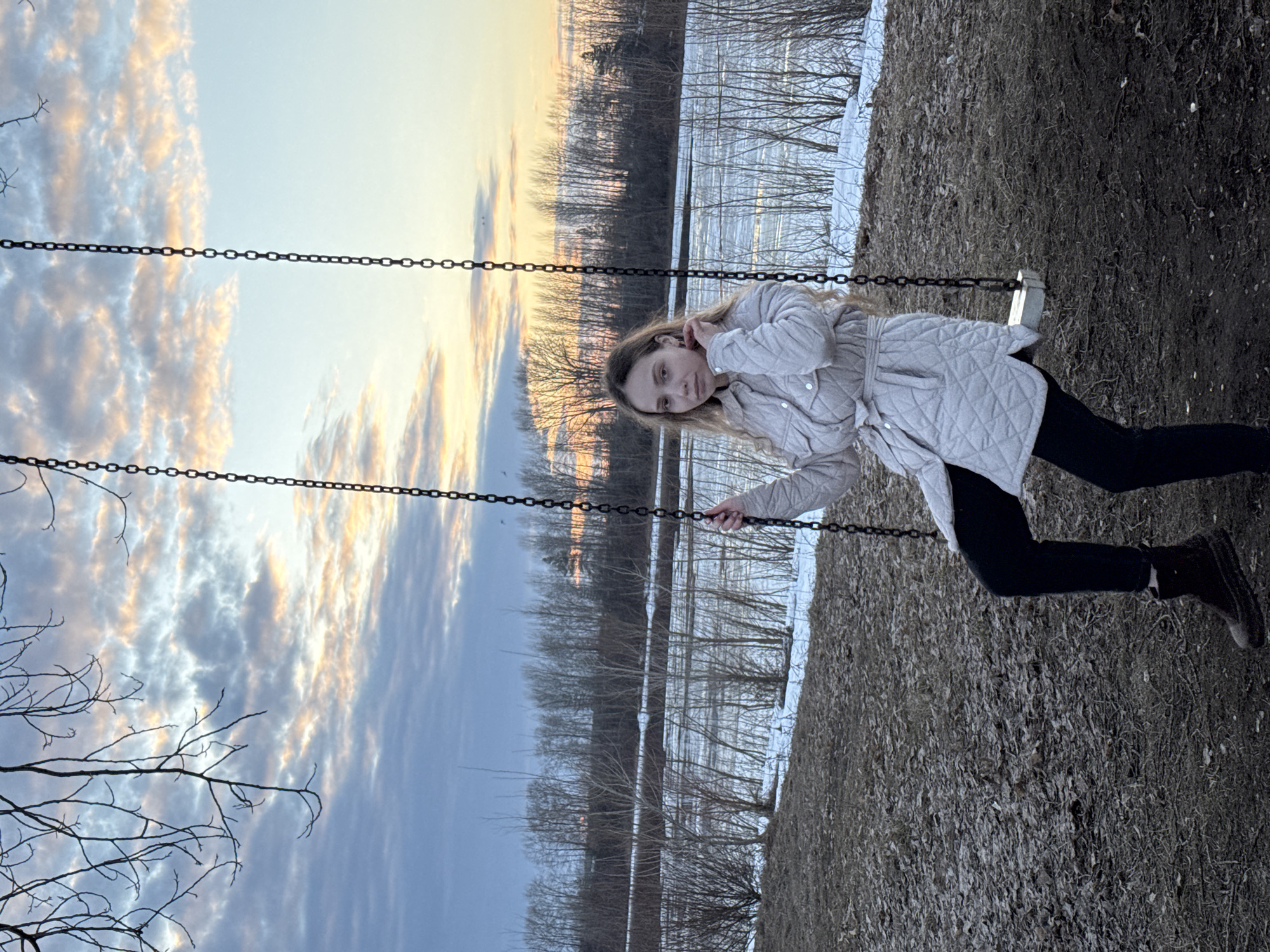 A young woman is on a swing in what appears to be a park. She wears a light gray quilted jacket, black leggings, and dark boots. The background shows a body of water, possibly a lake or river, and trees. The sky has a mix of clouds and clear blue with the setting sun creating a warm glow. The ground has leaves. The location is potentially Chaikovsky, Russia.