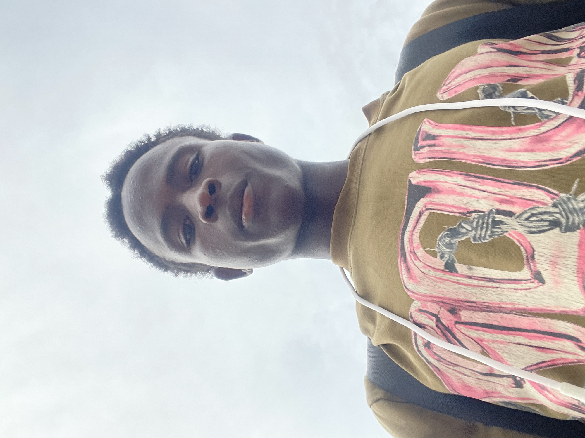 A low-angle, medium shot captures a young Black male from the chest up, facing forward. He has short, dark, curly hair and is looking directly at the camera with a neutral expression. He is wearing a tan sweatshirt with a large graphic design on the front featuring pink lettering intertwined with rope. A white drawstring is visible from the sweatshirt, and a dark strap, possibly from a backpack, is visible over his left shoulder. The background is a pale, overcast sky. The image appears to have been taken outdoors, possibly on a street or open area in Jos, Nigeria, given the provided context.