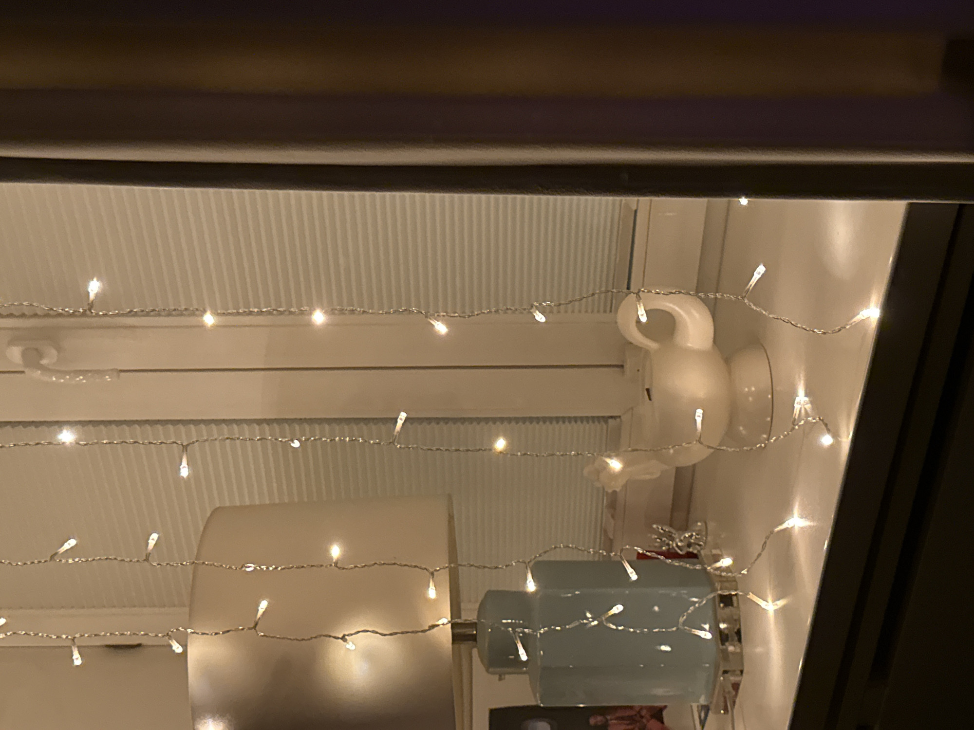 The image depicts a close-up view of a windowsill adorned with decorative lights. On the left, a pale blue ceramic lamp base with a cream-colored lampshade is visible. To its right, a white ceramic teapot shaped like a swan sits on the windowsill. Several strands of small, white LED lights hang vertically in front of a window with horizontal blinds. A dark brown curtain is visible on the right side of the frame. The overall scene is dimly lit, with the lights providing a warm, ambient glow. The location is identified as Unknown, Unknown.