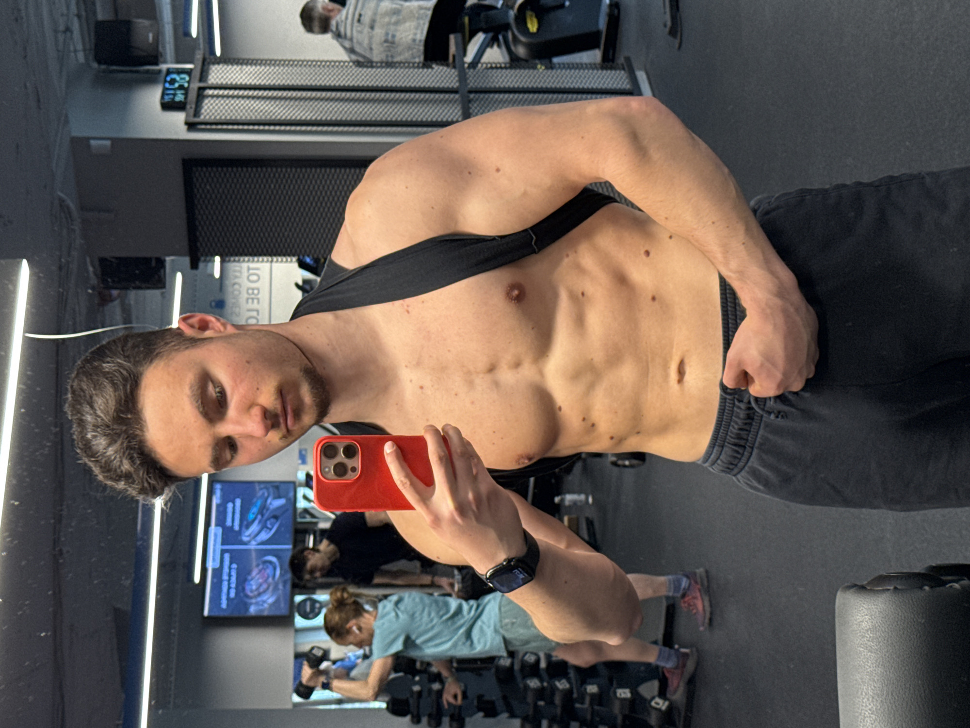 The image captures a mirror selfie taken by a young man inside a modern gym in Sofia, Bulgaria, likely in the morning given the visible digital clock. The main subject is a fit man with dark, wavy hair and light stubble, looking directly into the camera. He is shirtless, with a black athletic top pulled down around his shoulders, showcasing his muscular torso and arms. He wears dark shorts and a black smartwatch on his left wrist, and holds a red iPhone with a multi-lens camera to take the photo.
The gym environment features dark walls and flooring, illuminated by bright vertical LED strip lights. In the background, several other gym-goers are visible in reflection. To the upper right, a man in a light-colored, possibly plaid shirt is on a piece of cardio equipment, possibly a rower or elliptical. Another man in a black shirt is partially obscured behind the main subject's phone. To the lower left, a woman with light brown hair tied in a bun, wearing a light blue t-shirt and black shorts, is focused on lifting dumbbells. Various gym equipment, including weight racks and a padded bench, are also visible.
Notable details include a digital clock on the wall displaying "08:53" and "140". A large screen reflected in the mirror shows fragmented text like "TO BE TO" and "ITA COVEZ," along with images that might be workout diagrams or app interfaces. The overall atmosphere suggests a focused workout session in a well-equipped fitness center.