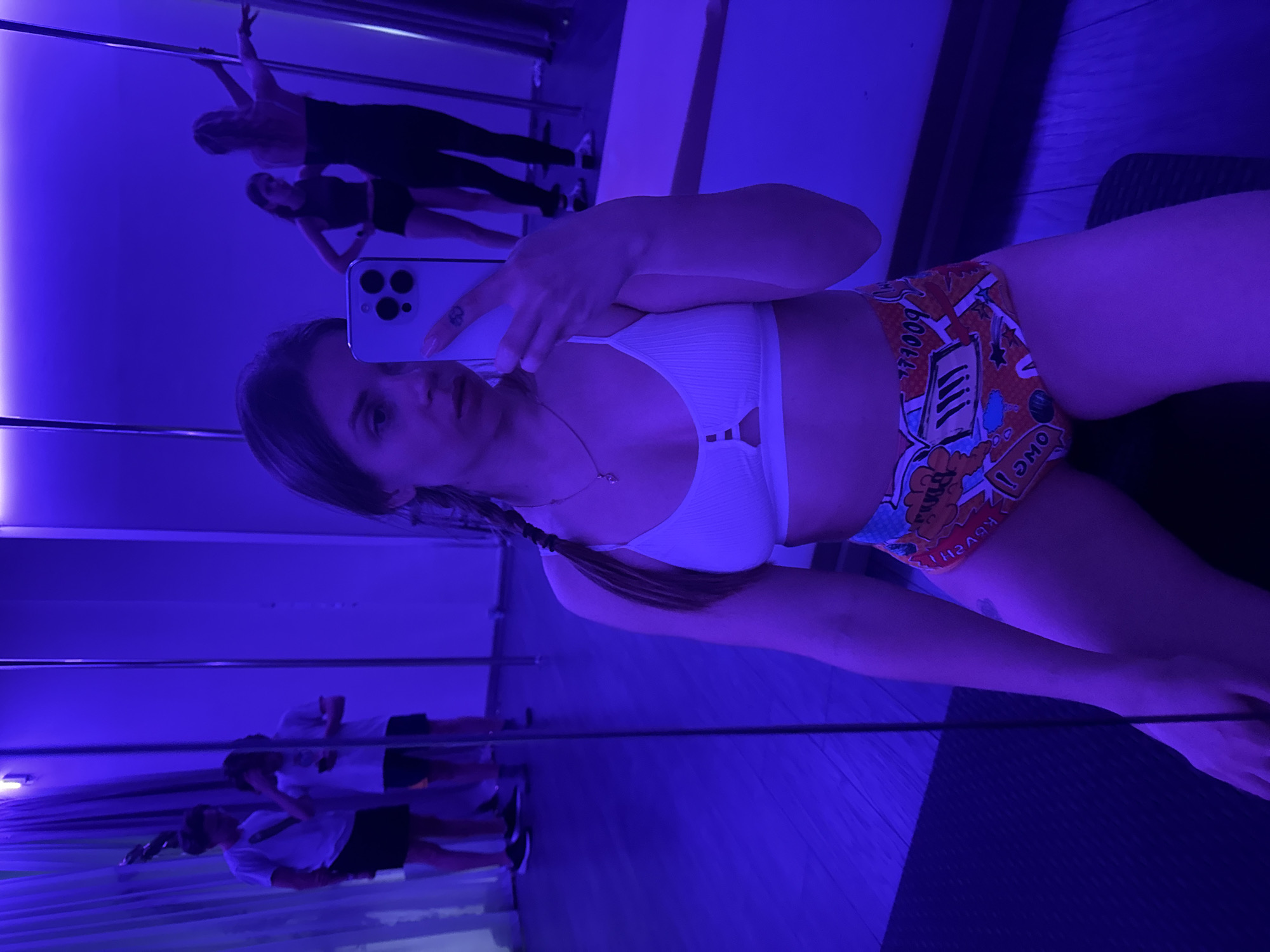 The image appears to be a selfie taken in a dance studio or fitness space in Batumi, Georgia. A woman with braided hair is holding a smartphone, likely an iPhone, taking a picture of herself in a mirror. She is wearing a white sports bra and printed shorts. The room is lit with a blue light and has a mirror covering at least one wall, in which other people are visible, including some engaging in pole dancing. Vertical poles are also visible in the reflection. The floor appears to be wooden, possibly covered in a black mat in the foreground.