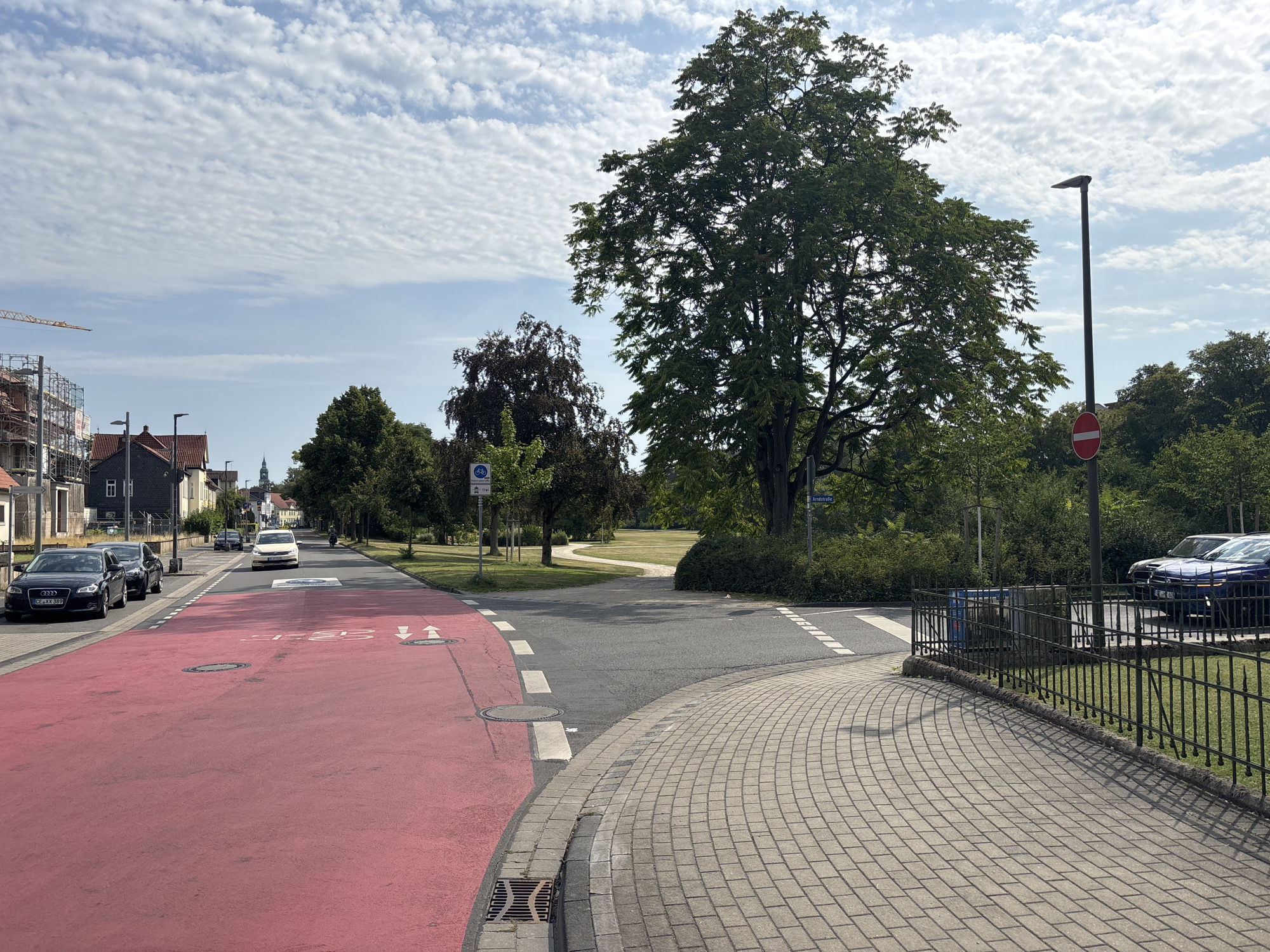 The image shows a street scene in Celle, Germany, on a cloudy day. In the foreground, a red-colored bike lane curves to the right, bordered by a sidewalk made of paving stones. A black metal fence runs along the edge of the sidewalk. Beyond the bike lane and sidewalk, a grey asphalt road continues, with a pedestrian crossing marked by white lines. Several cars are parked on the left side of the road, including a black Audi with a German license plate. A white taxi is driving on the road. In the background, there are trees, buildings, and a street lamp. Construction scaffolding is visible on a building to the left. The sky is filled with wispy clouds. A street sign with the name "Arndtstraße" is visible on the right.