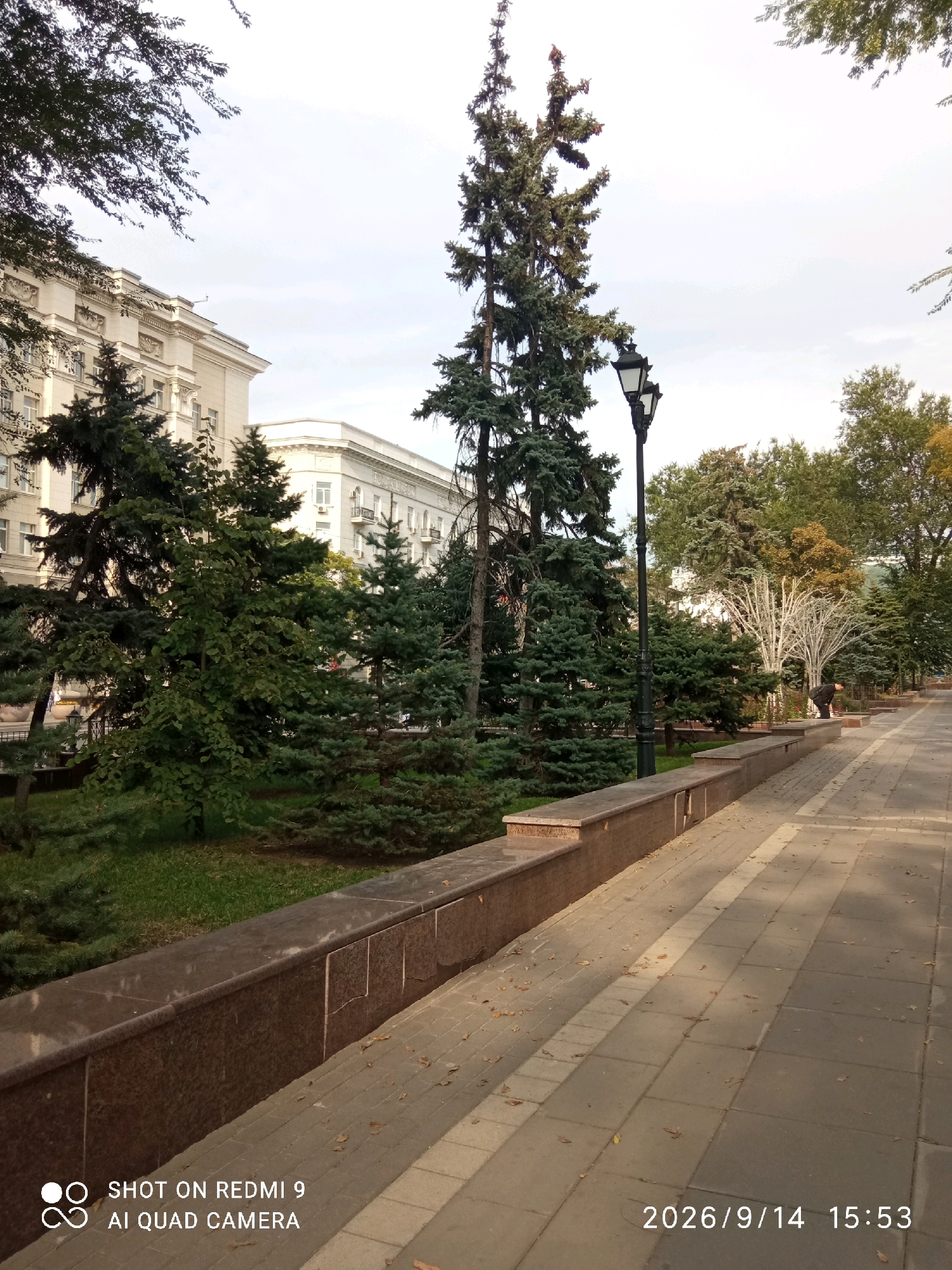 A paved pedestrian walkway lined with trees and a stone embankment stretches into the distance under an overcast sky. The walkway is made of rectangular pavers and scattered fallen leaves. A tall, black street lamp with two lights stands on the left side of the walkway, behind a dense cluster of evergreen trees. A low, dark stone embankment with granite detailing runs along the left side of the walkway, separating it from a grassy area with more trees. In the background, a large, ornate, multi-story building with many windows and decorative architectural elements is visible. Further down the walkway, a person in dark clothing is bending over. The timestamp "2026/9/14 15:53" and "SHOT ON REDMI 9 AI QUAD CAMERA" are visible in the bottom right and bottom left corners, respectively. The scene appears to be a park or a boulevard within an urban setting. Based on the timestamp, the location is likely Rostov-on-Don, Russia.