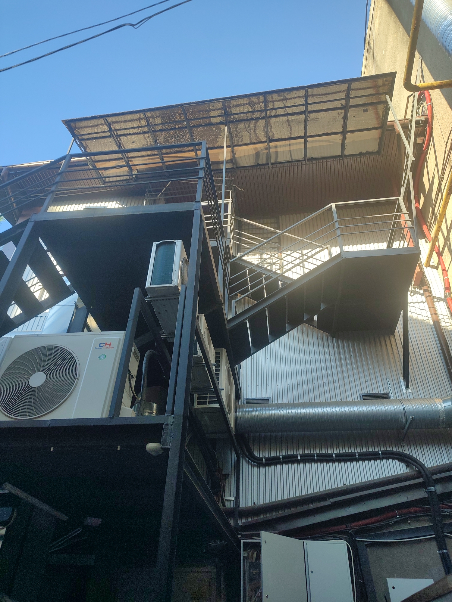 The image is a low-angle, outdoor photograph showing the exterior of a building with multiple levels of outdoor metal staircases and platforms. The building's facade appears to be corrugated metal, with various pipes and ventilation systems attached. Several outdoor air conditioning units are visible on the platforms. The sky above is clear and blue. The photograph was taken in Chernivtsi, Ukraine.